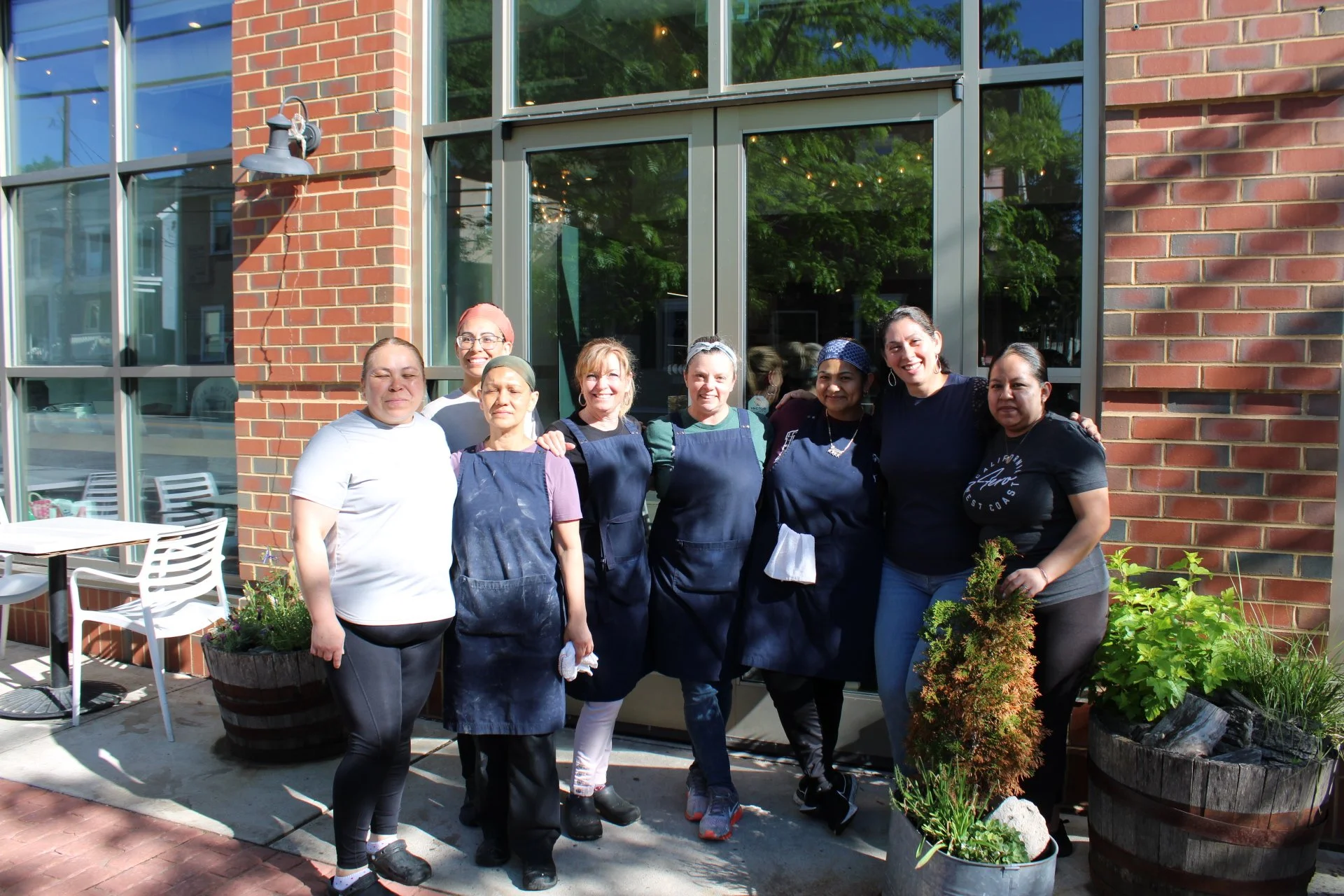 Group of women standing outside near a brick building with large glass windows, some wearing aprons, posing for a photo.