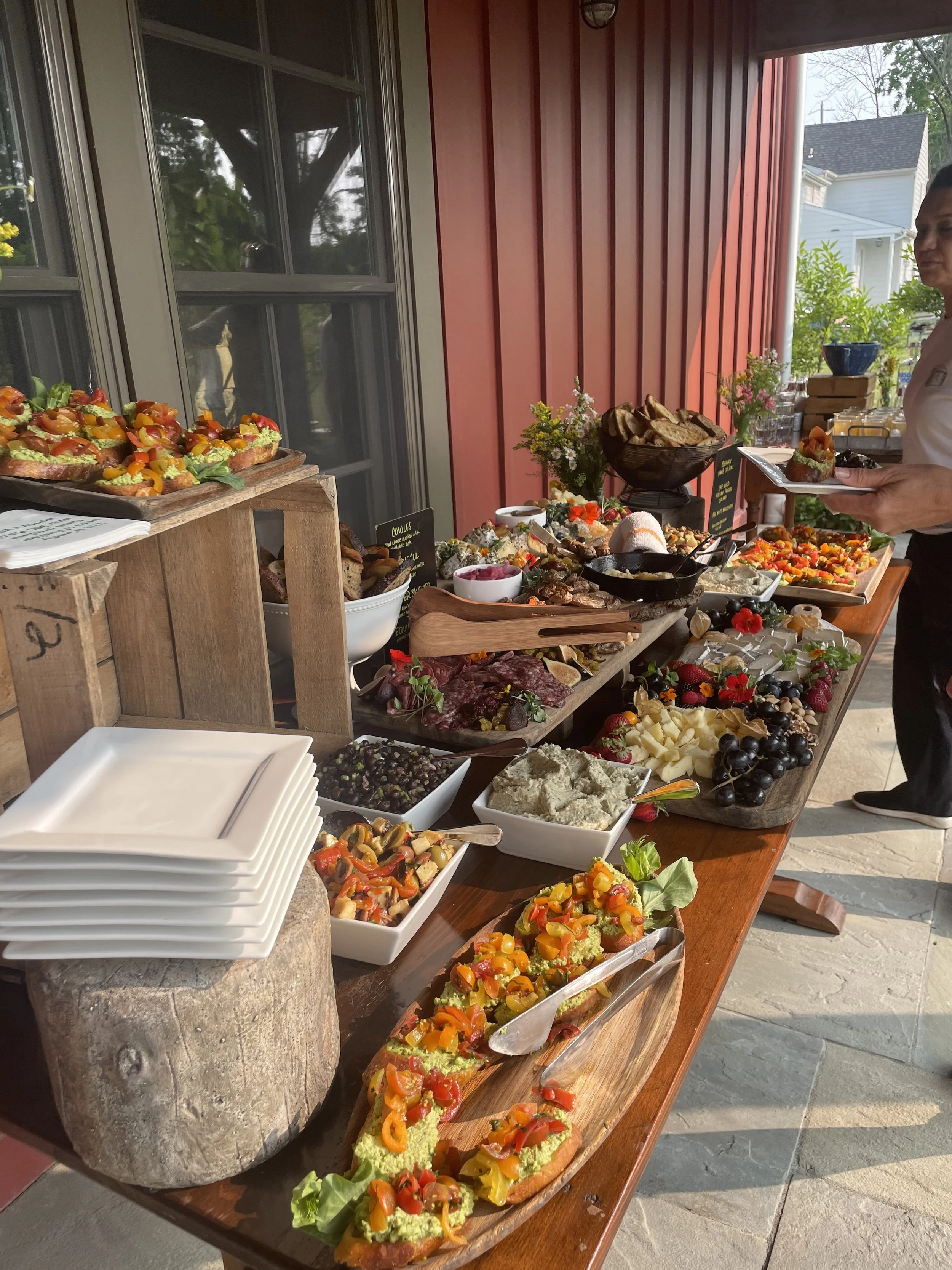 A buffet table with various colorful dishes, including decorated bread or bruschetta, cheeses, fruits, salads, and spreads, set outside on a porch with a red wall and house in the background.