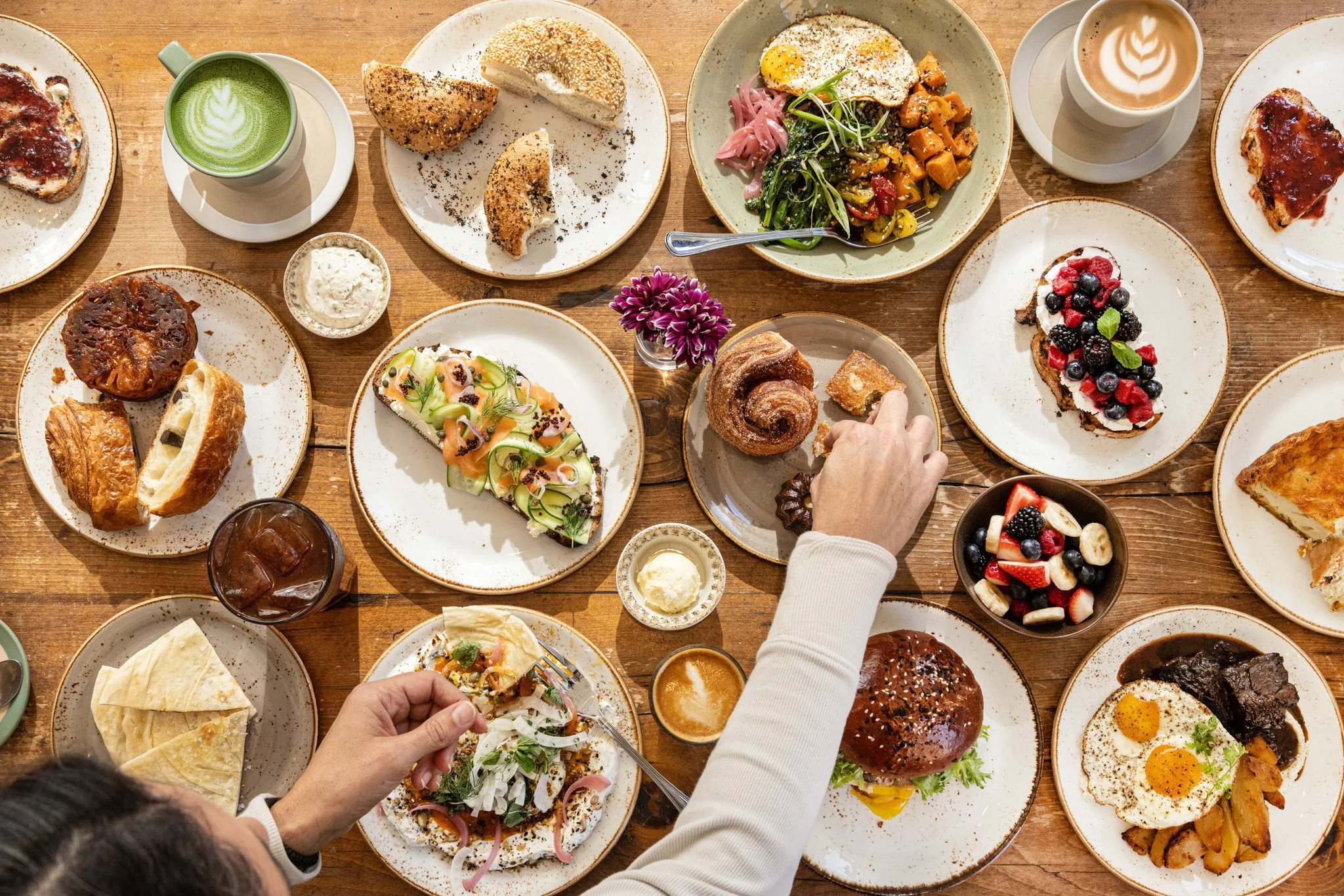 A top-down view of a table filled with various breakfast dishes, including pancakes, toast, salads, a burger, a fruit tart, baked goods, coffee, and juice, with a person's hand reaching for a pastry.