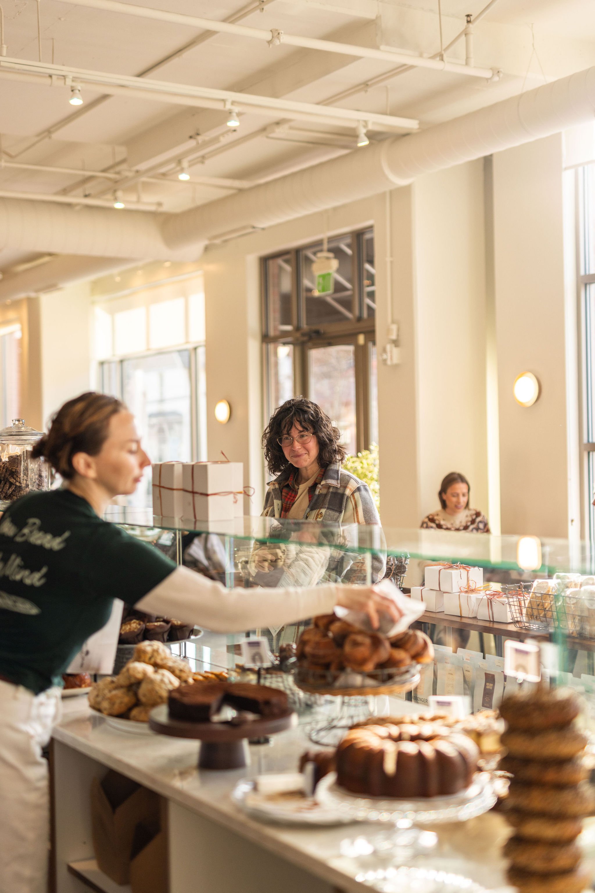 Inside a bakery, a woman with short hair and eyeglasses stands behind the counter, smiling at a customer. The customer, a woman with curly hair and glasses, is reaching for pastries. Various baked goods, including cakes, cookies, and muffins, are displayed on the counter. Sunlight streams through large windows, illuminating the warm, inviting atmosphere.