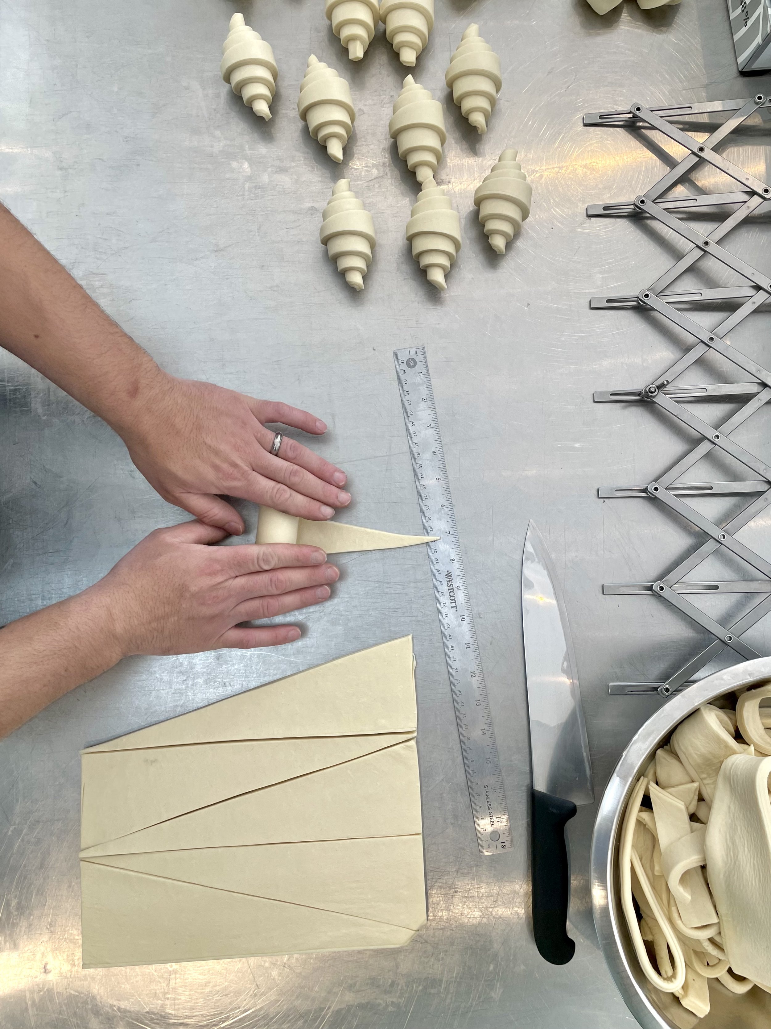 Person cutting rolled dough into strips on a stainless steel work surface surrounded by uncooked pastry spirals, a ruler, a knife, and a bowl of cut dough.