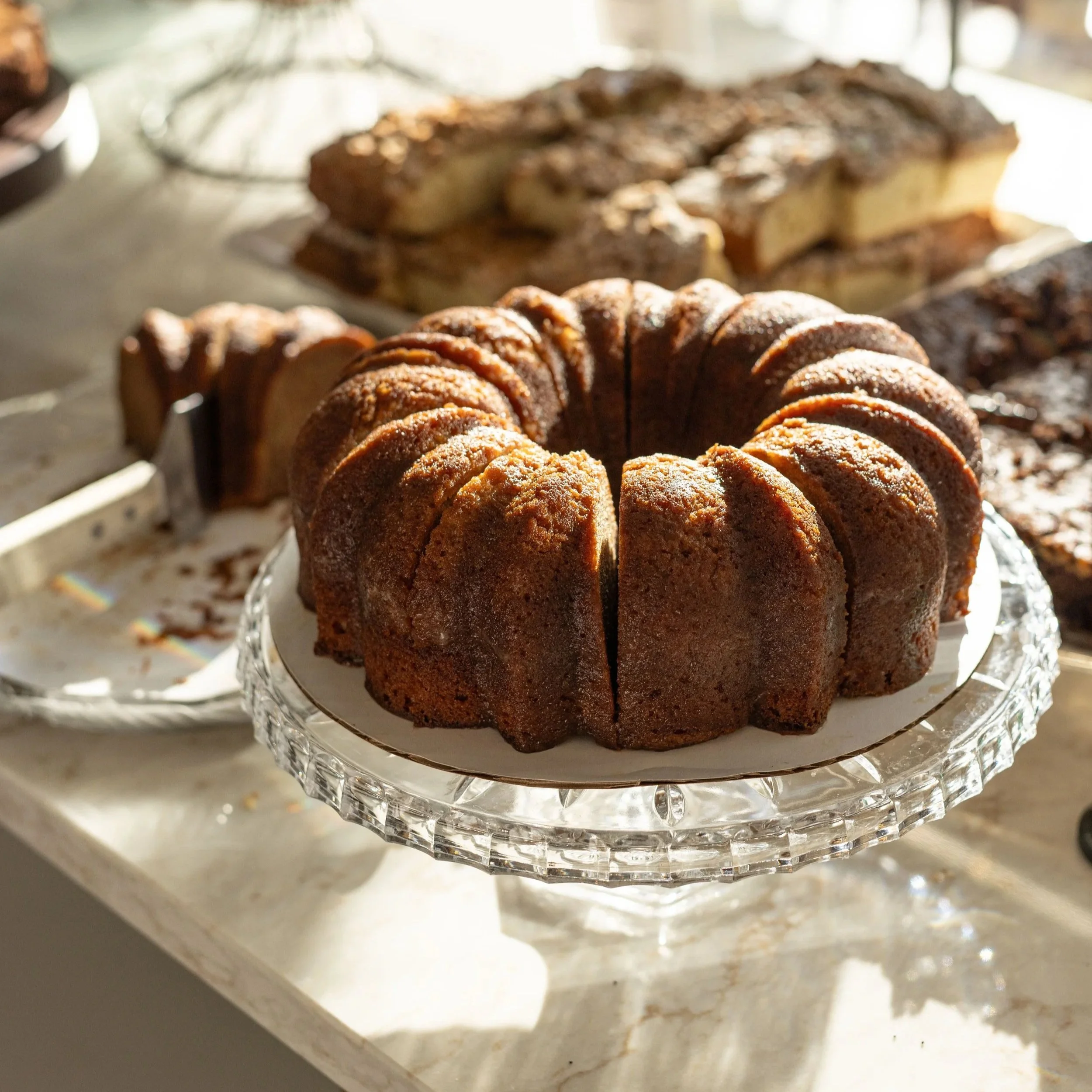 Bundt cake dusted with powdered sugar on a glass cake stand with other baked goods in the background.