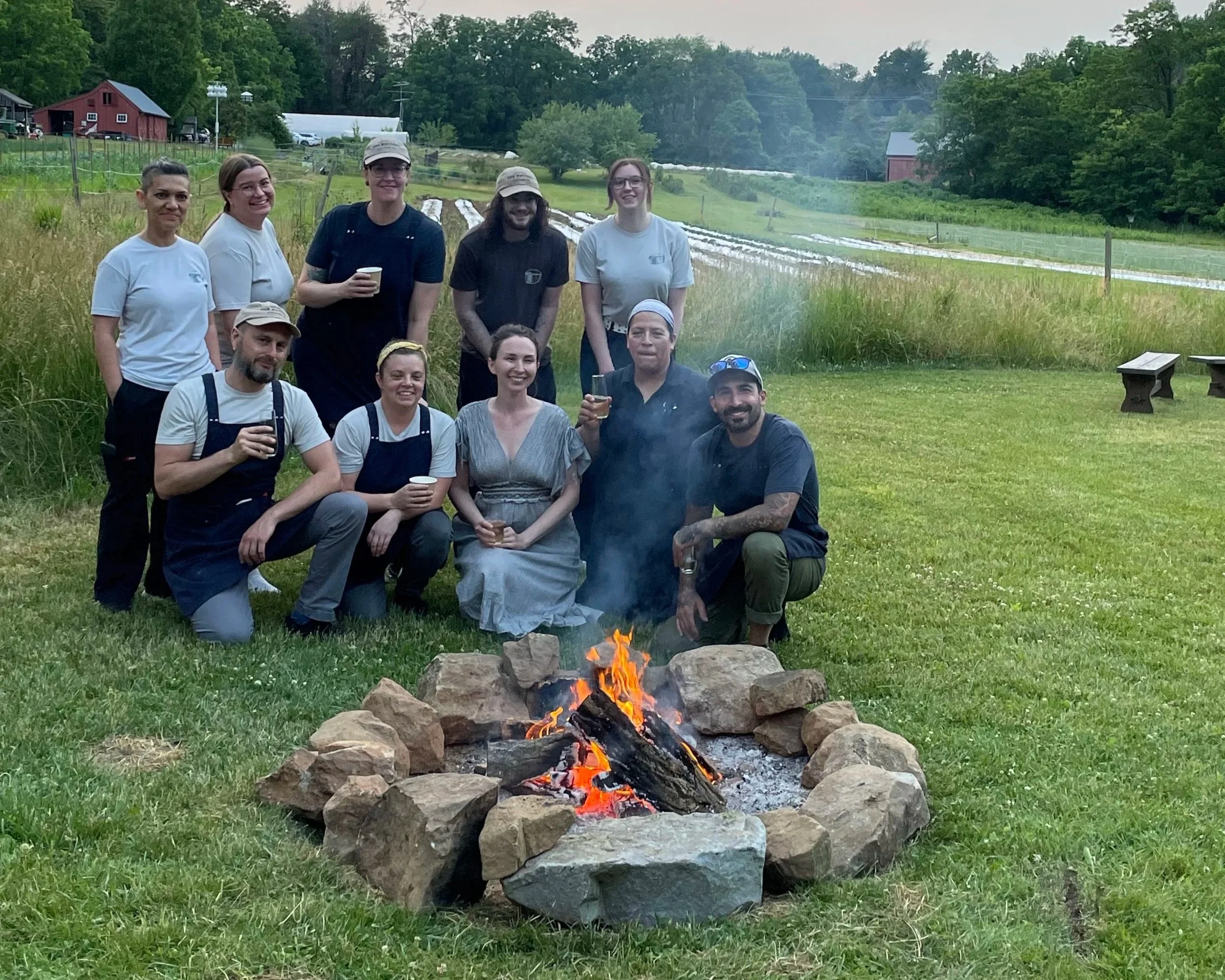 A group of ten people gathered outdoors around a campfire with a rural landscape background, some holding cups, smiling, and posing for the photo.