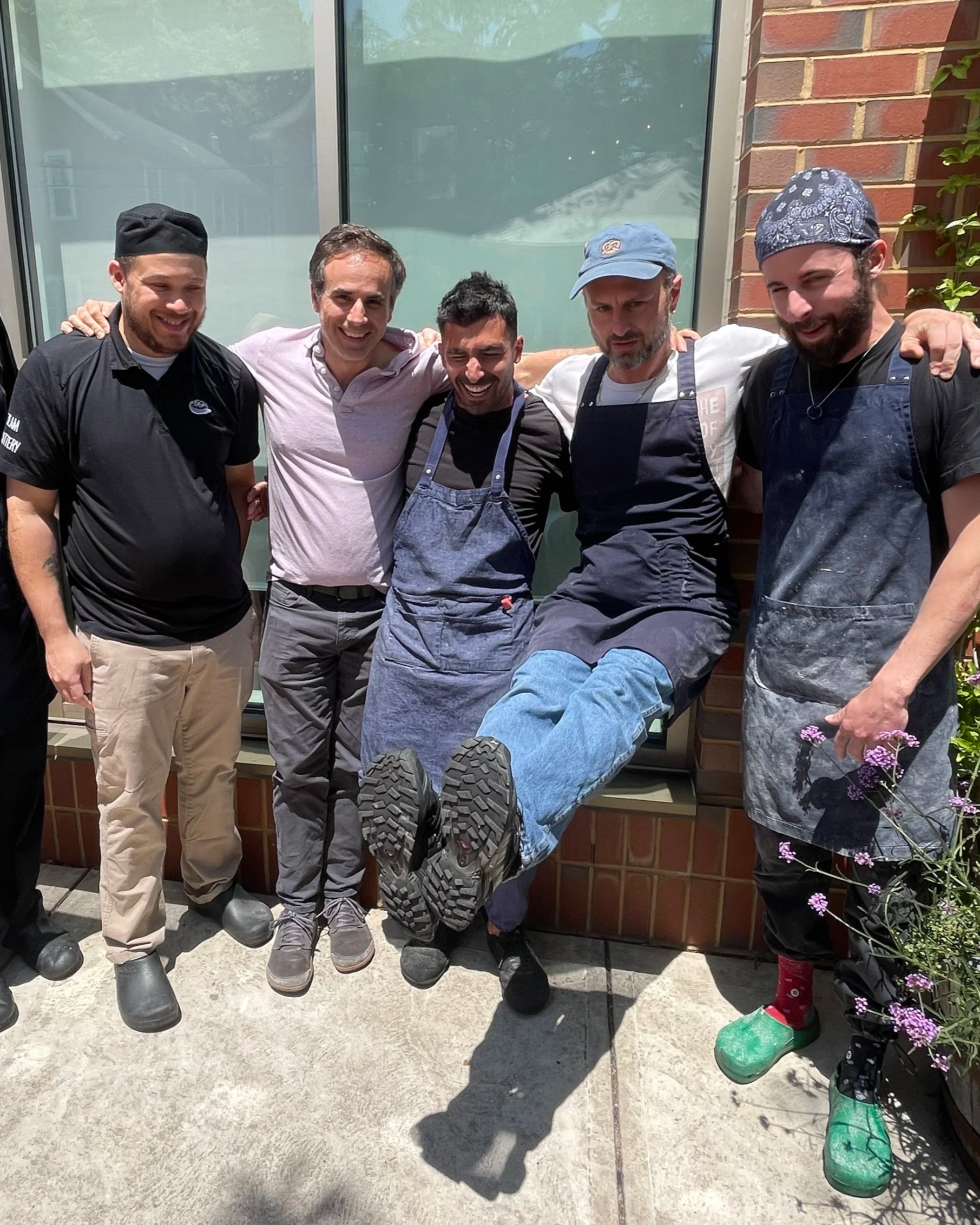 Group of five men standing together outdoors, with one man being lifted and supported by the others, all smiling and looking at the camera.