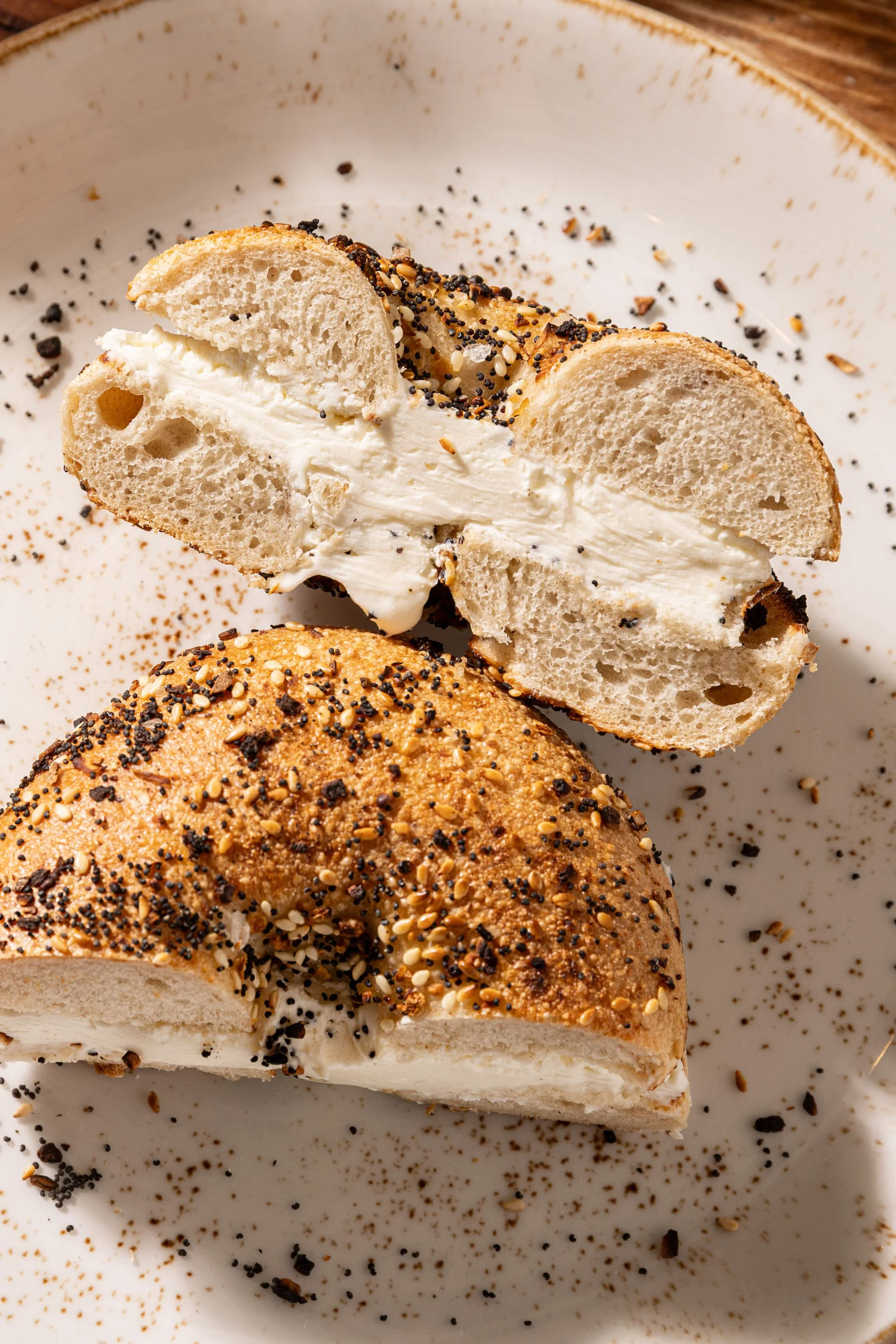 A bagel sliced in half, filled with cream cheese, topped with poppy seeds and sesame seeds, placed on a white plate