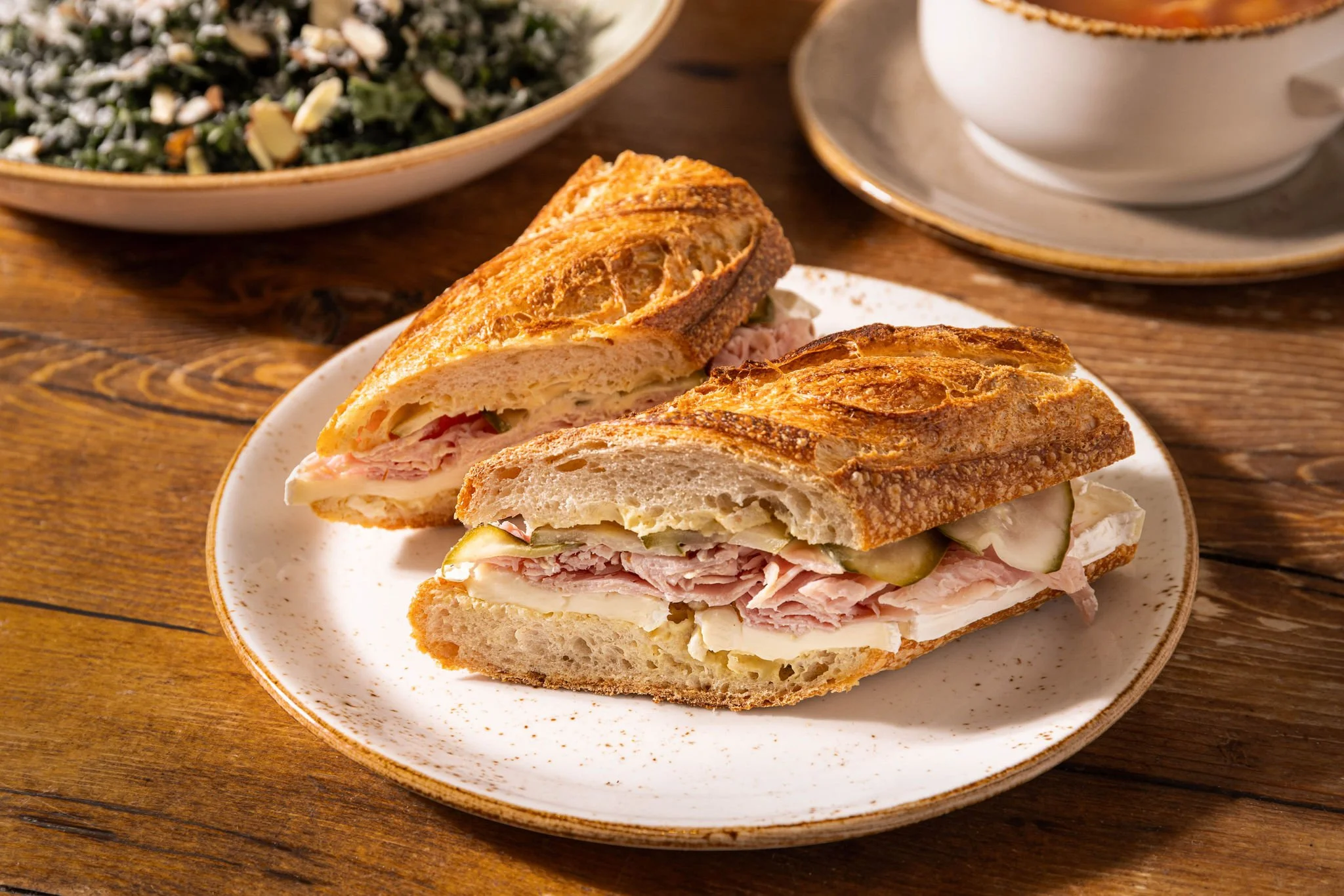 A toasted sandwich with layers of ham, cheese, and pickles inside a sliced French baguette on a white speckled plate. A bowl of salad and a cup of coffee sit on a wooden table in the background.