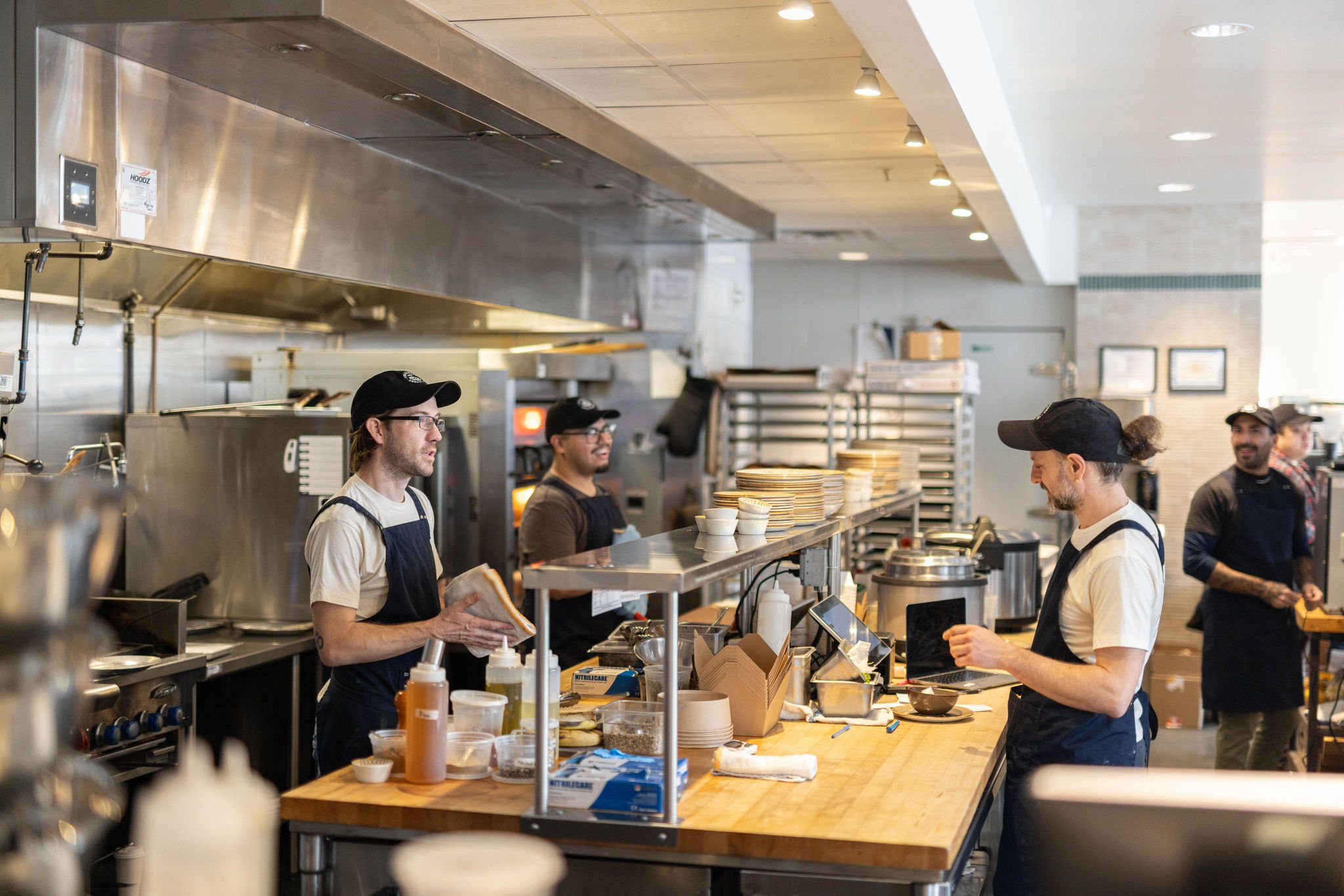 Four restaurant workers wearing aprons and hats working behind the kitchen counter, with stacks of plates and kitchen utensils visible.
