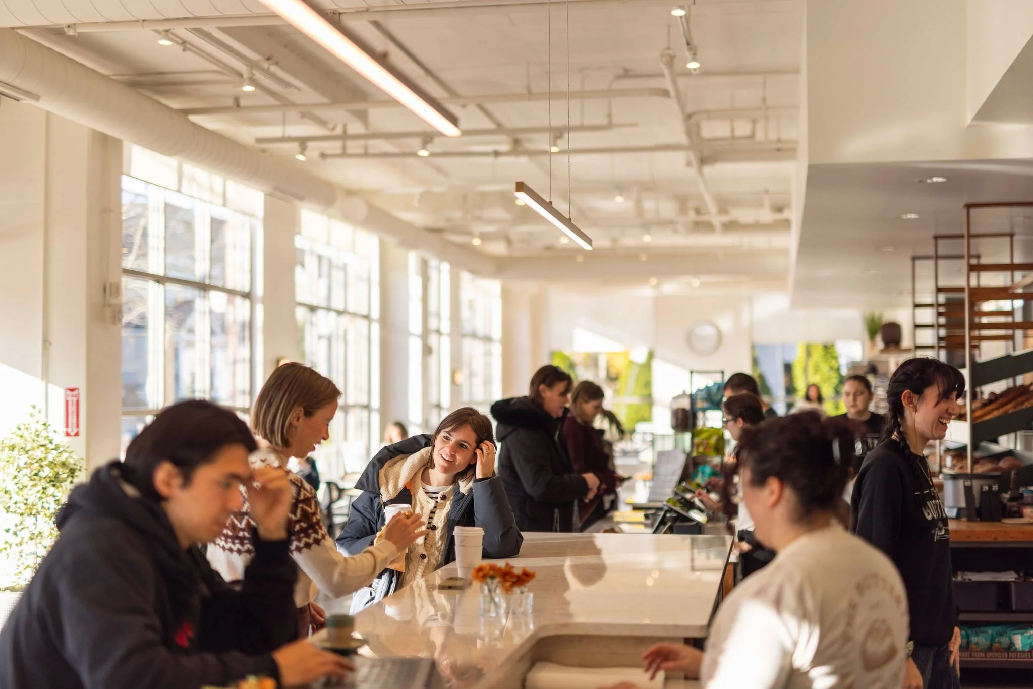People ordering and socializing at a cafe counter inside a bright, modern cafe with large windows and indoor plants.