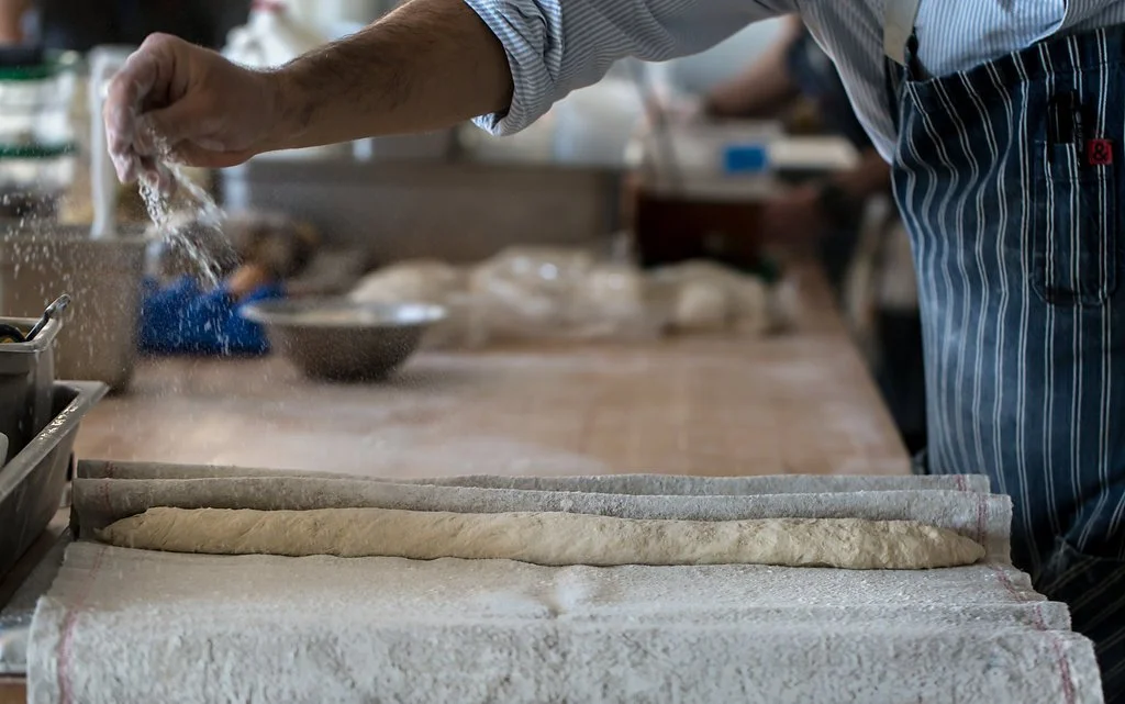 Person sprinkling flour on rolled dough in a bakery kitchen.