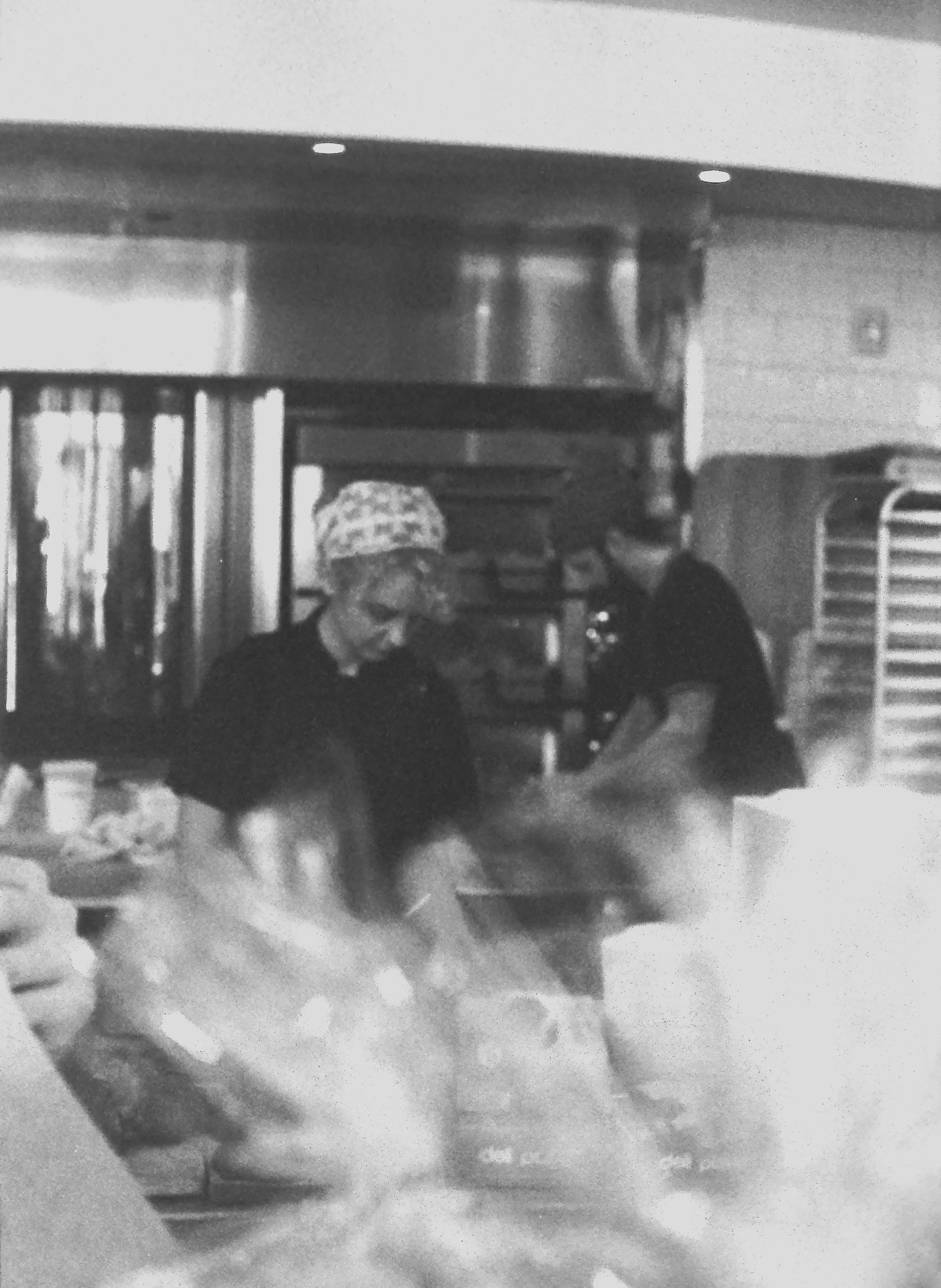 Two women working in a commercial kitchen, wearing hair coverings and preparing food.