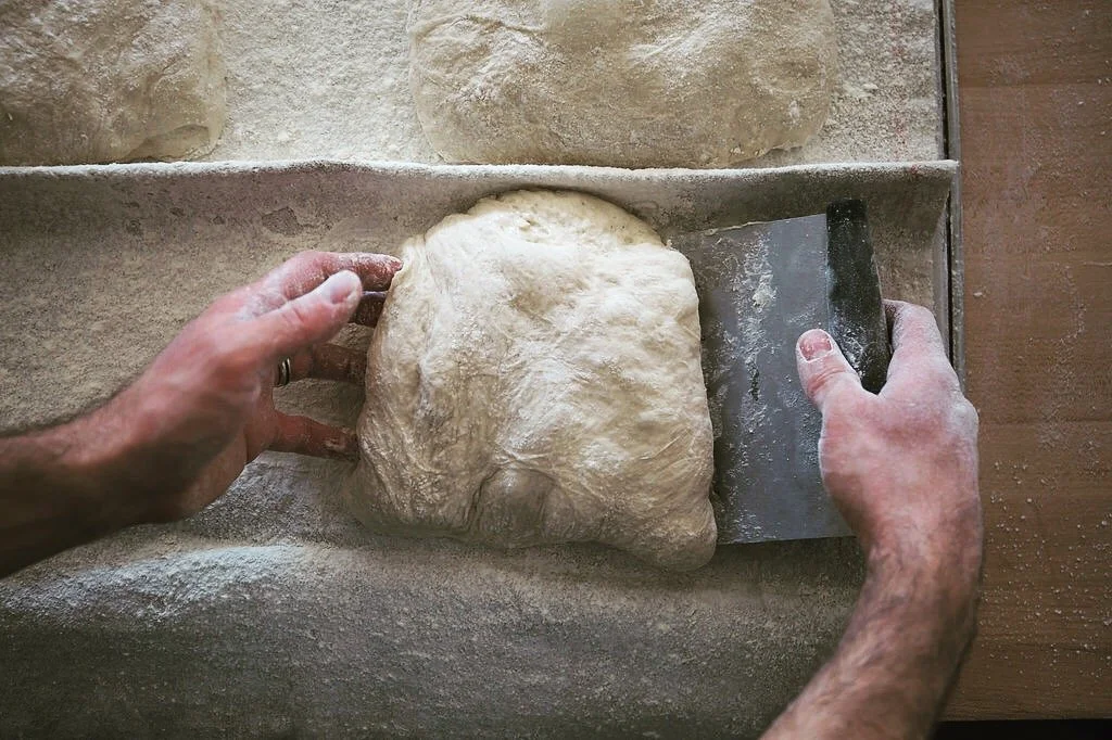 A person kneading dough with a bench scraper on a floured stone surface.