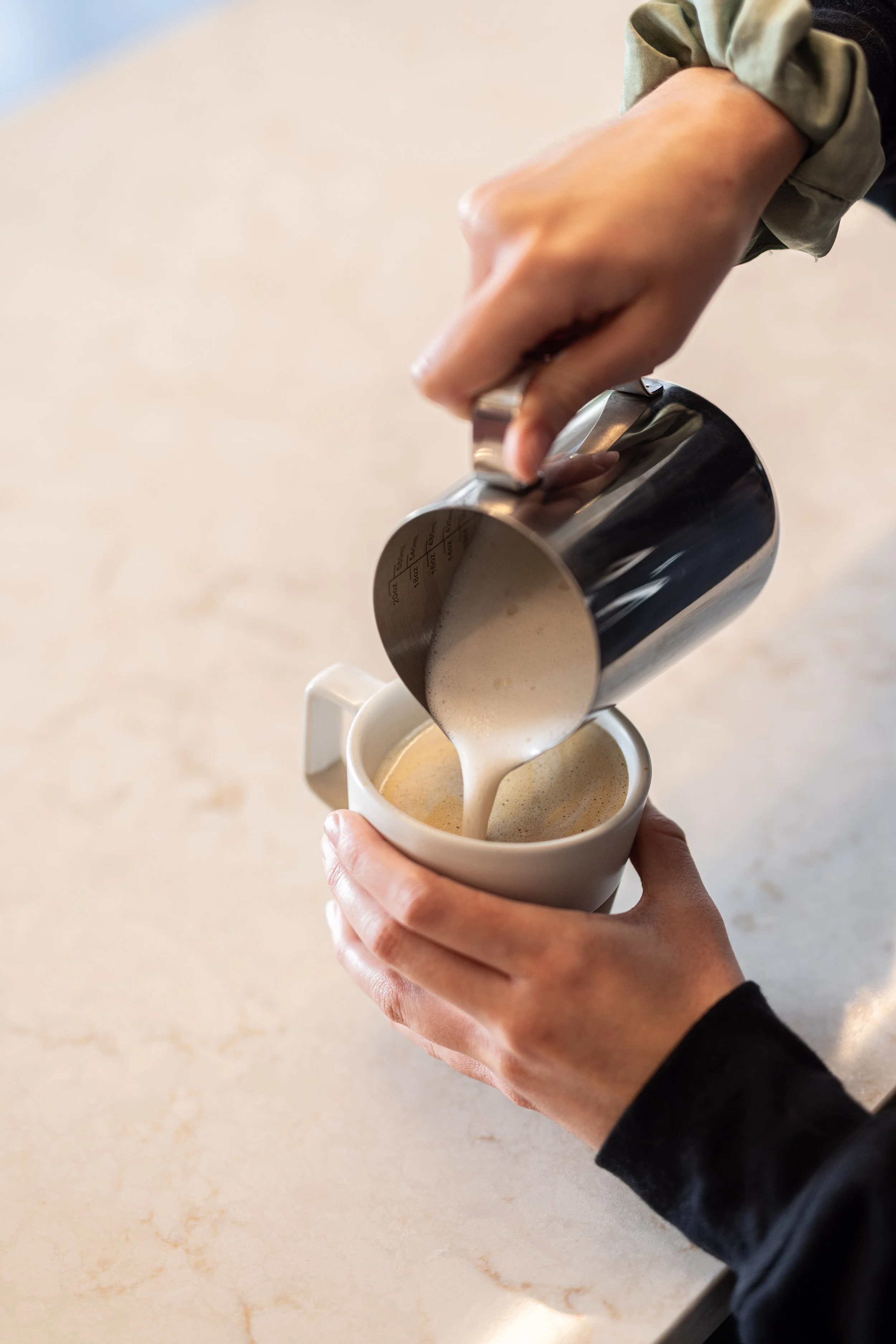Person pouring frothed cream or milk into a white coffee mug from a stainless steel measuring pitcher.