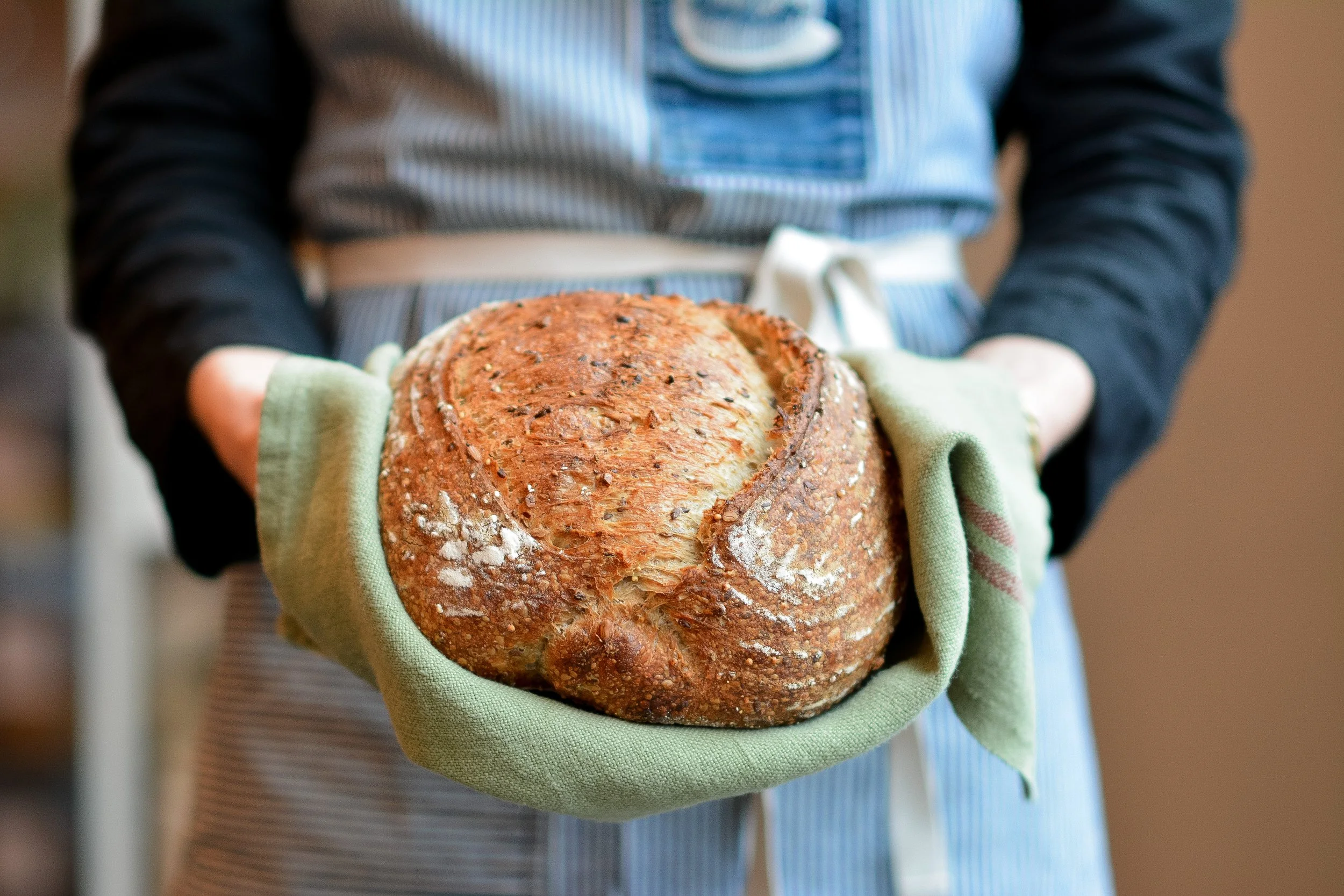 Person holding a freshly baked round loaf of bread with a crusty, textured surface, in green oven mitts, with an apron and striped shirt.