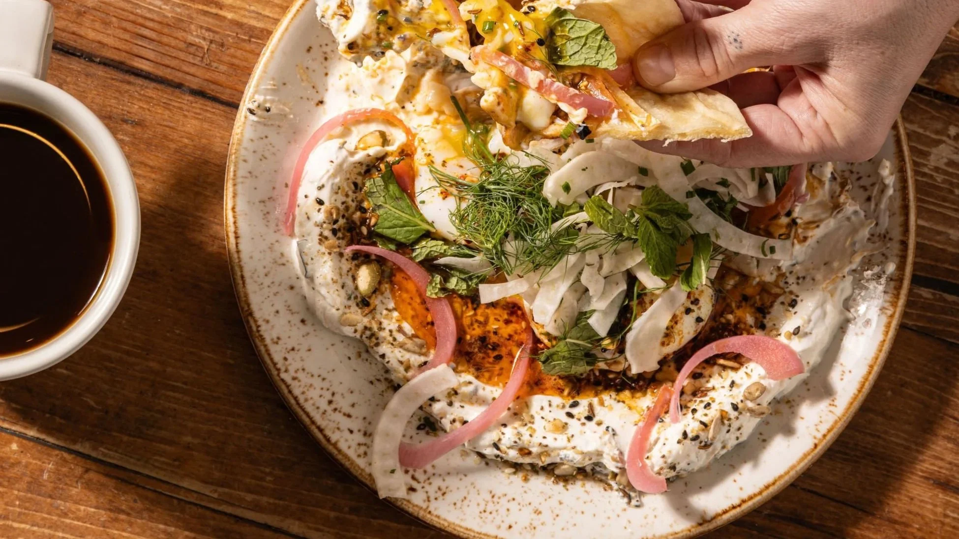 A plate of food topped with herbs, onions, and seeds, next to a cup of coffee on a wooden table.