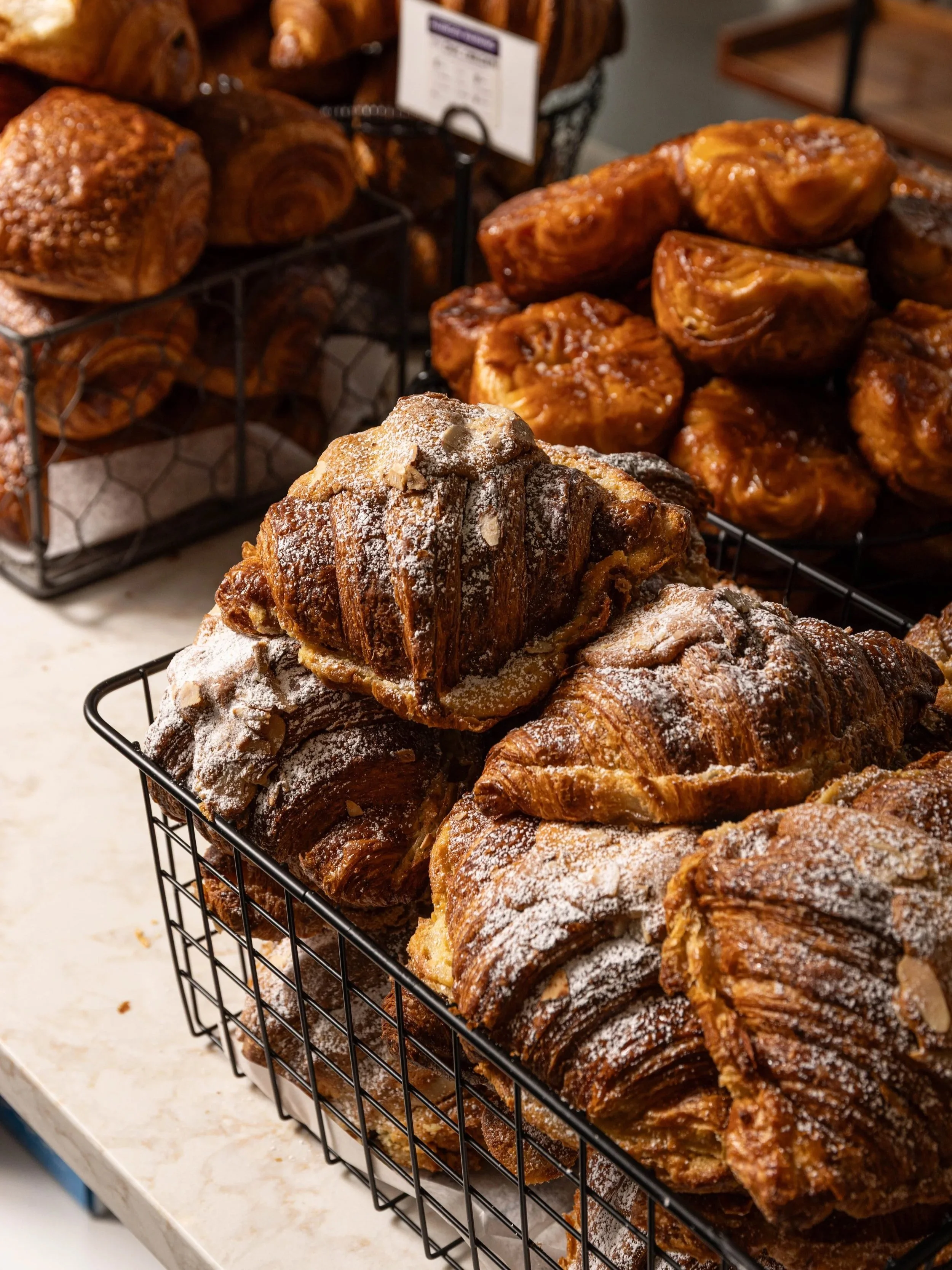 A black wire basket filled with assorted croissants dusted with powdered sugar, with more baked goods visible in the background.