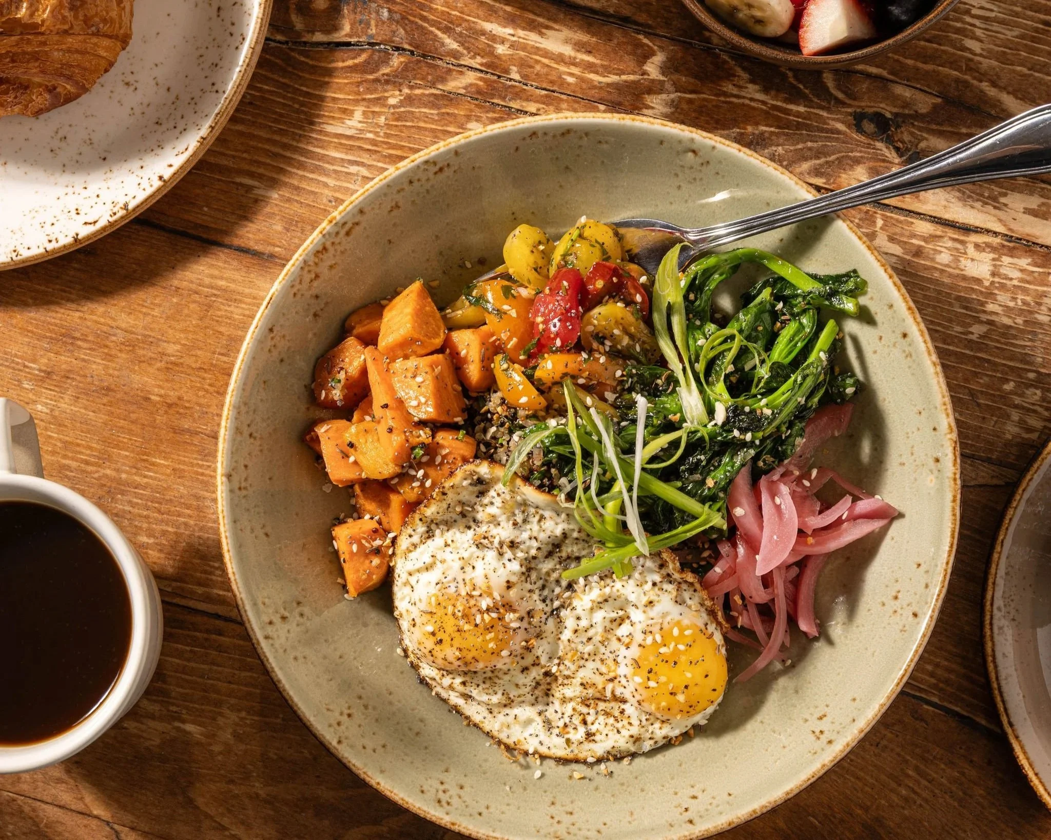 A breakfast dish in a bowl featuring sweet potatoes, cherry tomatoes, greens, and pickled onions, topped with two fried eggs, served on a rustic wooden table.