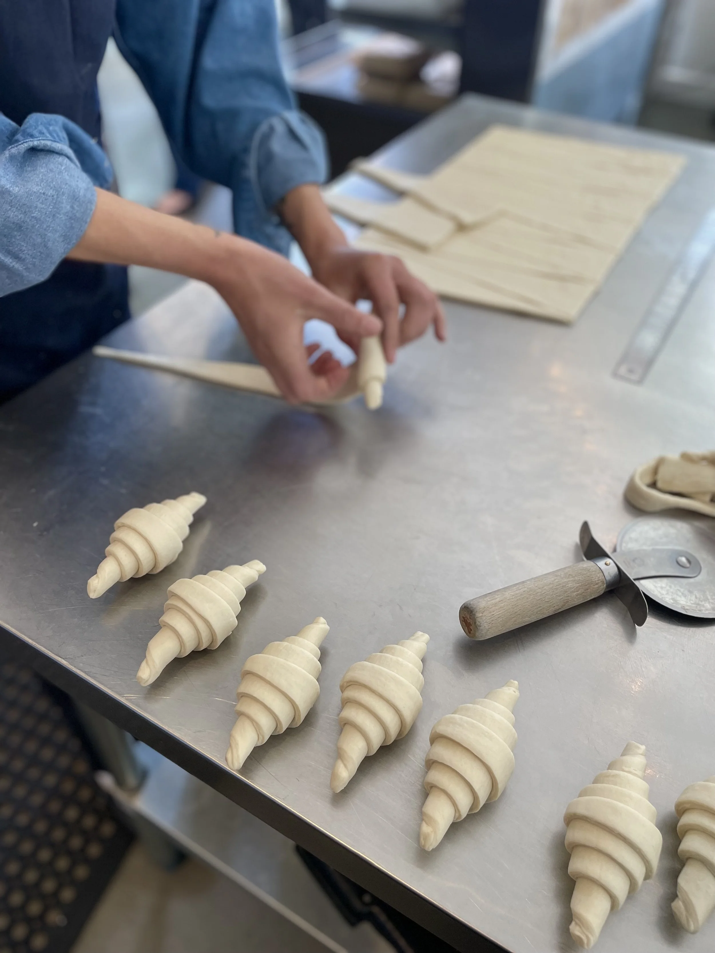 People preparing croissants on a stainless steel countertop with ready-to-bake croissants lined up, baking tools, and dough in the background.