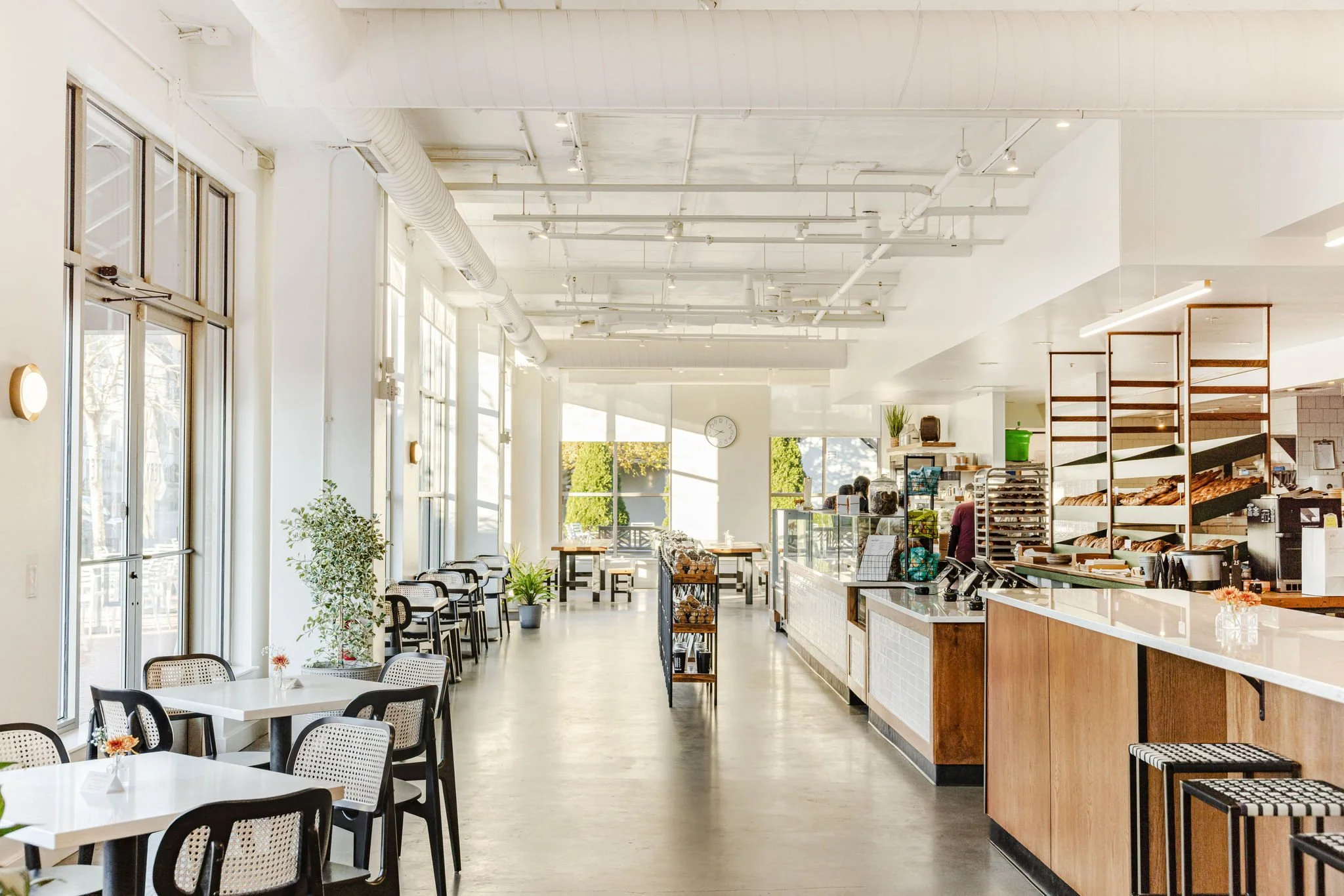 Bright, modern cafe with white walls, large windows, and minimal decor. Contains tables and chairs on the left, a counter on the right, a display of baked goods, and a sunny outdoor view.