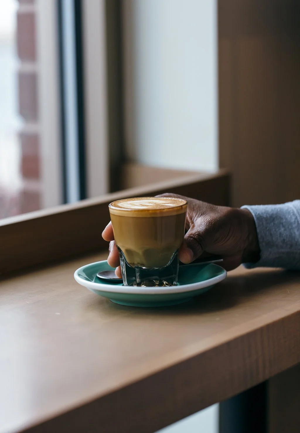 A person holding a latte in a glass on a saucer with a spoon, sitting by a window in a cozy cafe.
