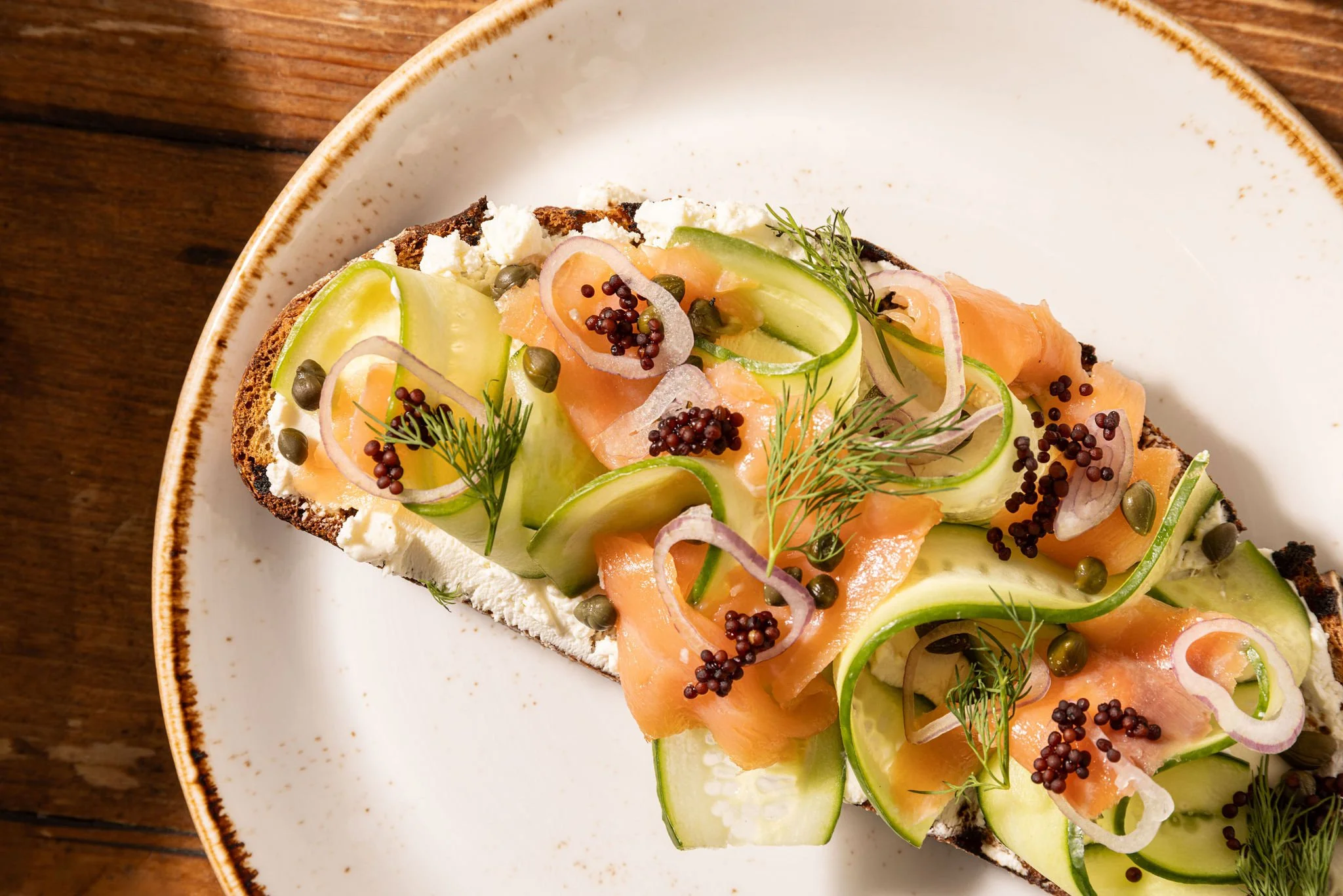 Open-faced sandwich with smoked salmon, cucumber slices, red onion, dill, capers, and mustard seeds on toasted bread with cream cheese, served on a white plate with a wooden background.