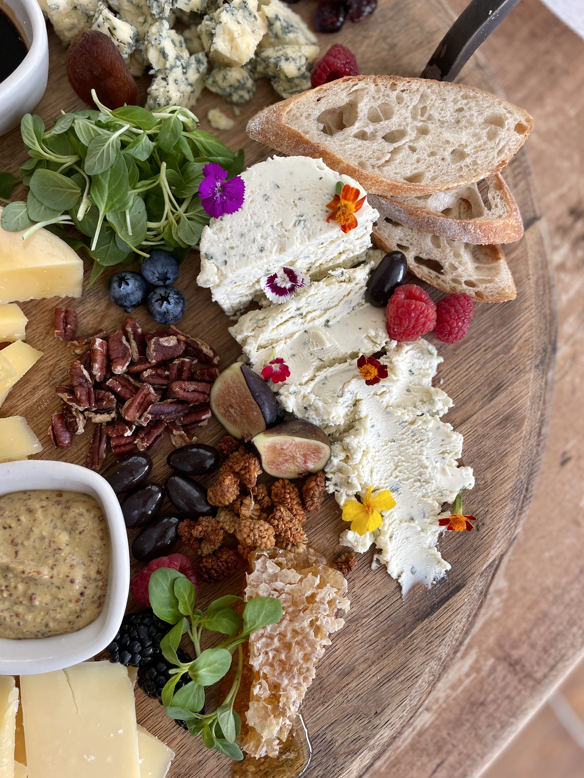 A cheese platter featuring various cheeses, fresh berries, nuts, honey, bread slices, and edible flowers on a wooden board.