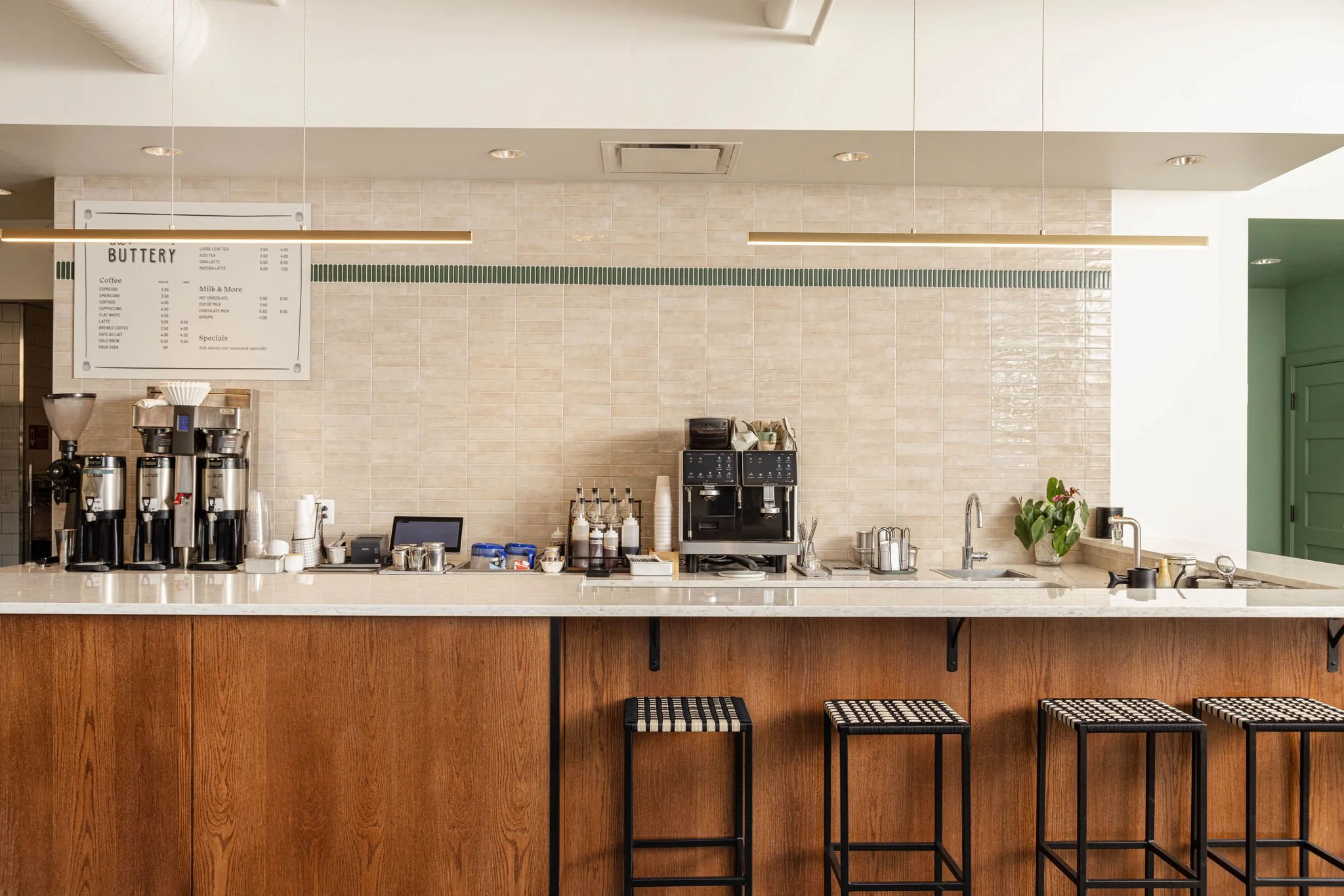 A modern coffee shop counter with three bar stools, coffee machines, syrups, and a menu board.