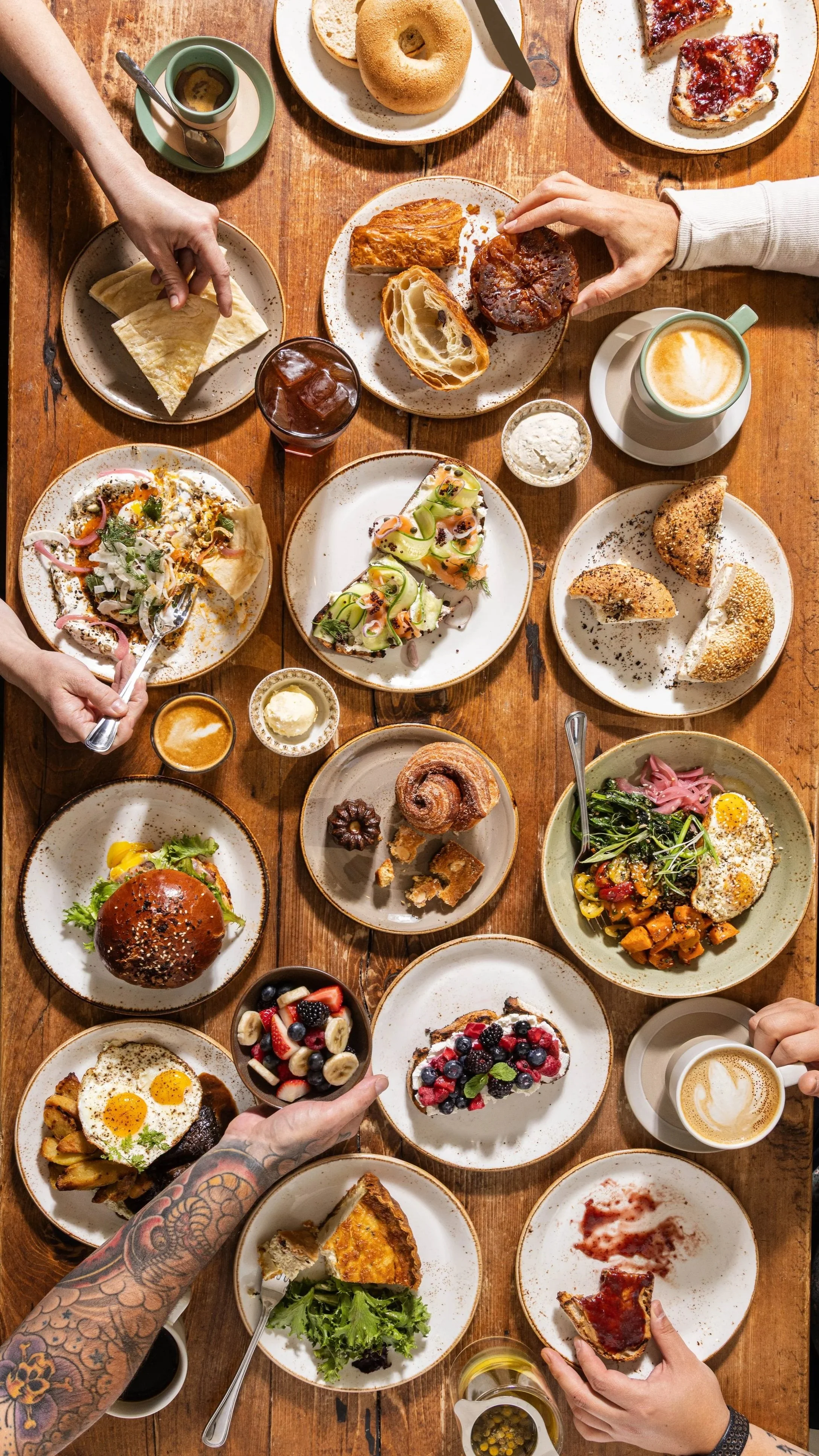 A table filled with various breakfast foods, including bagels, pizza slices, salads, sandwiches, fruit, pastries, coffee, and drinks, with multiple people reaching for the food.
