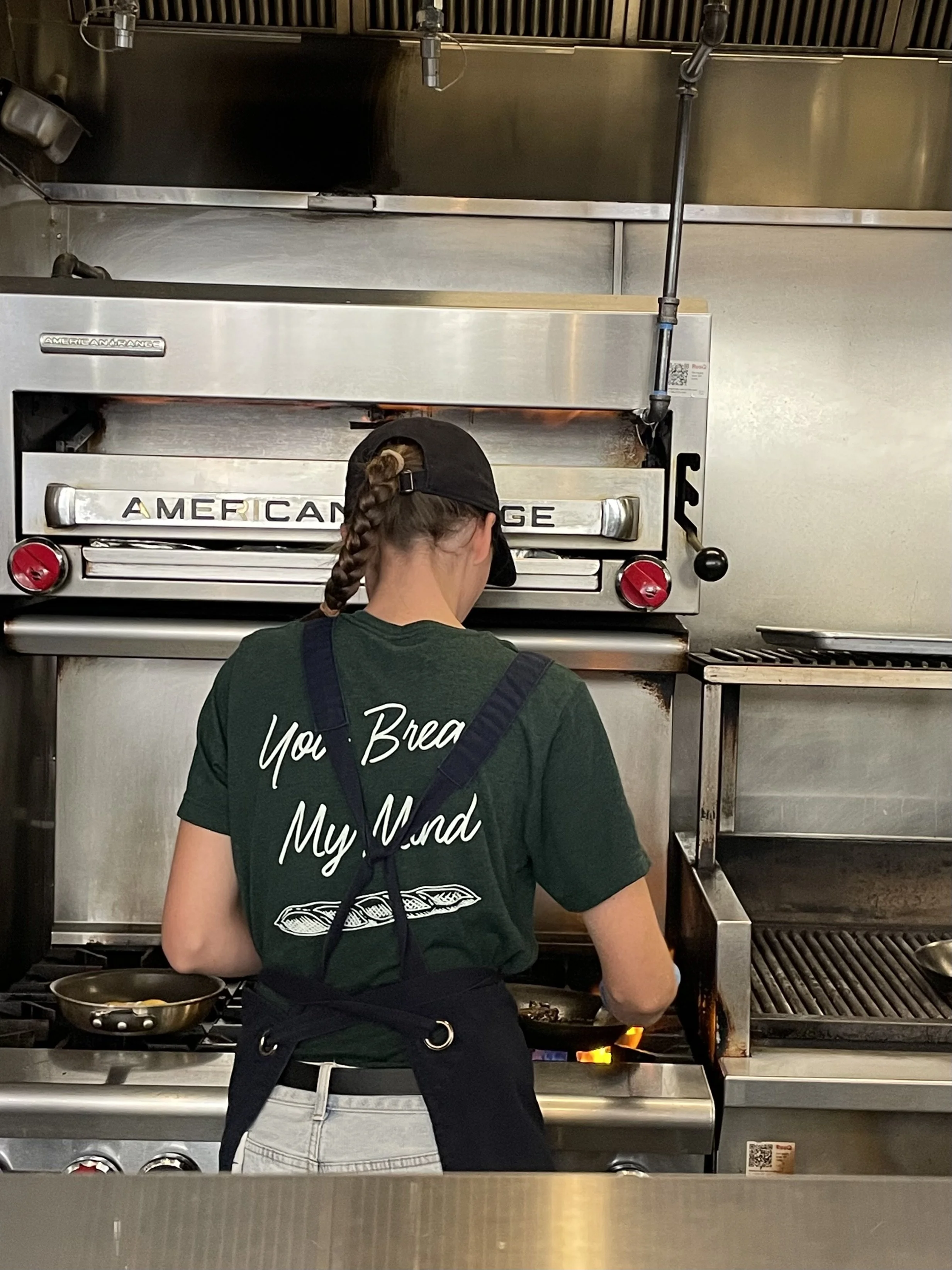 A woman in a restaurant kitchen wearing a green shirt with the text "You Bread My Mind" and an image of a baguette, and a black apron.