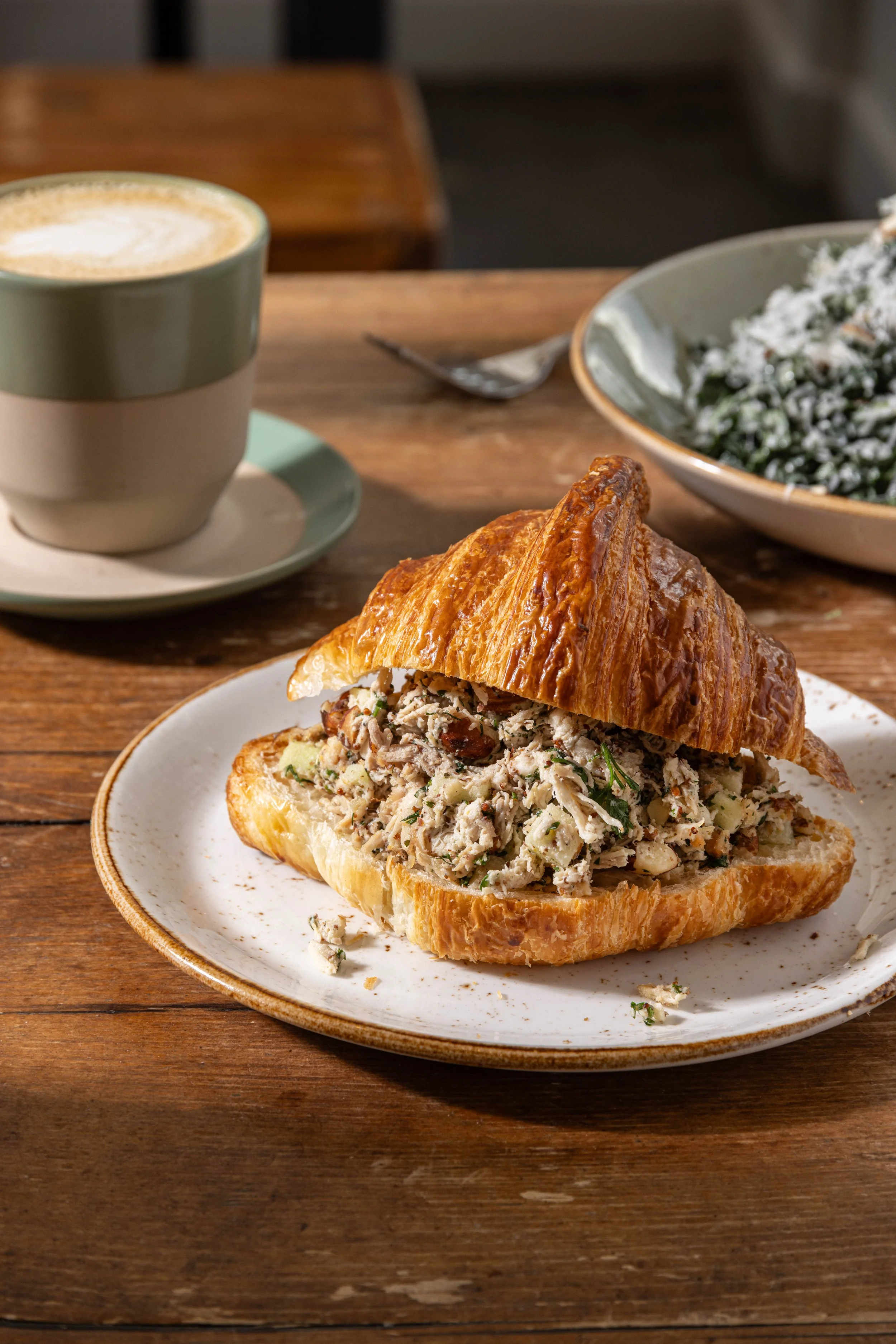 A croissant sandwich filled with chicken salad on a white plate, with a cup of coffee and a bowl of salad in the background on a wooden table.