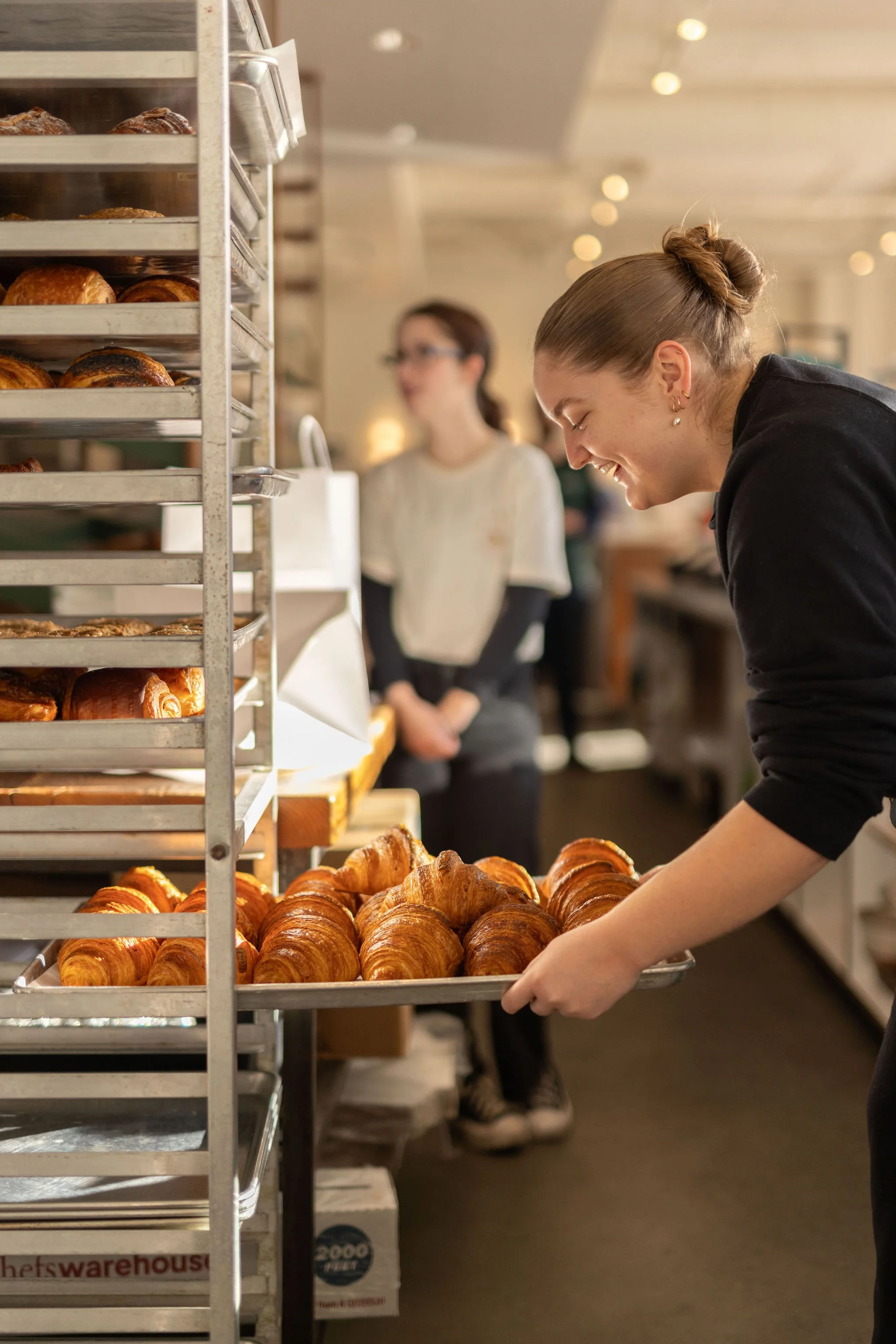 A woman smiling as she puts a tray of croissants into a bakery display.