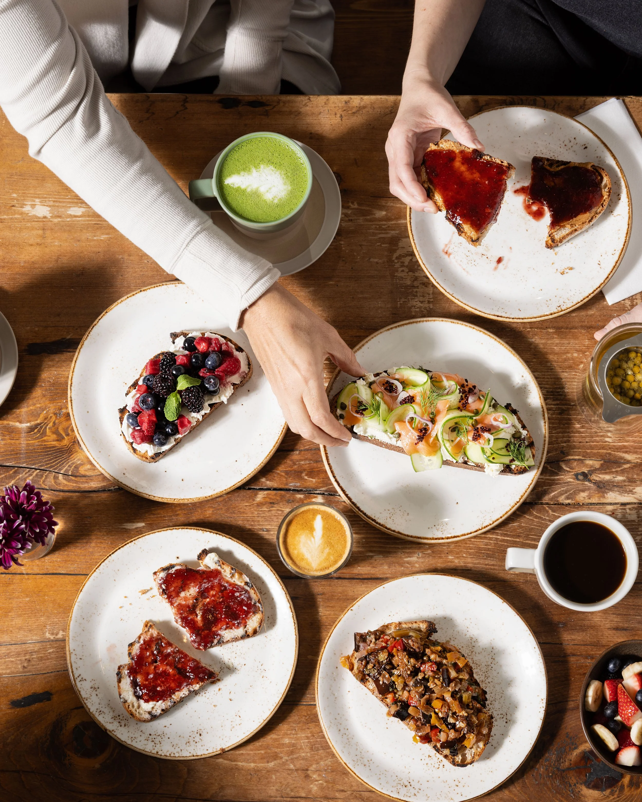 Top-down view of a wooden table with various food and drinks including fruit-topped bread, toast with jam, a green matcha latte, a hot coffee, a small espresso shot, and a small jar of passion fruit, with two people reaching for the food.