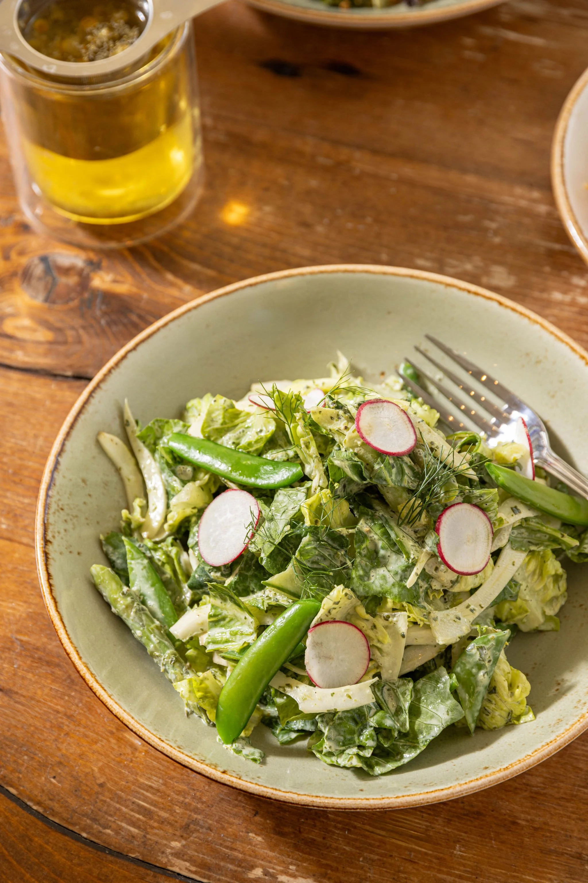 Fresh green salad with radish slices and snap peas in a beige bowl on a wooden table, with a fork and a glass of beer in the background.