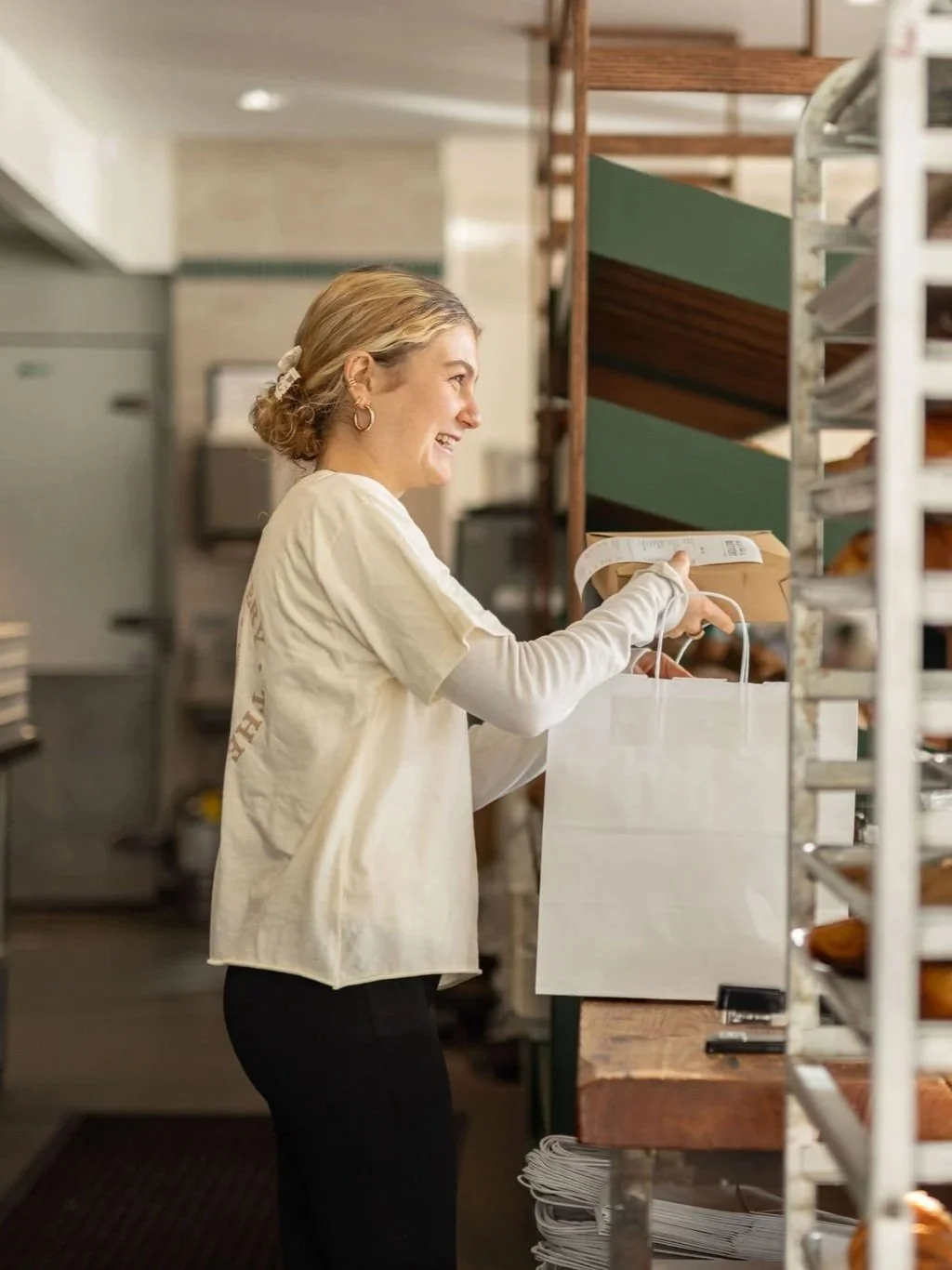 A woman with blonde hair tied back, wearing a beige T-shirt and black pants, smiling as she looks at a grocery bag she is holding in a bakery or grocery store aisle.
