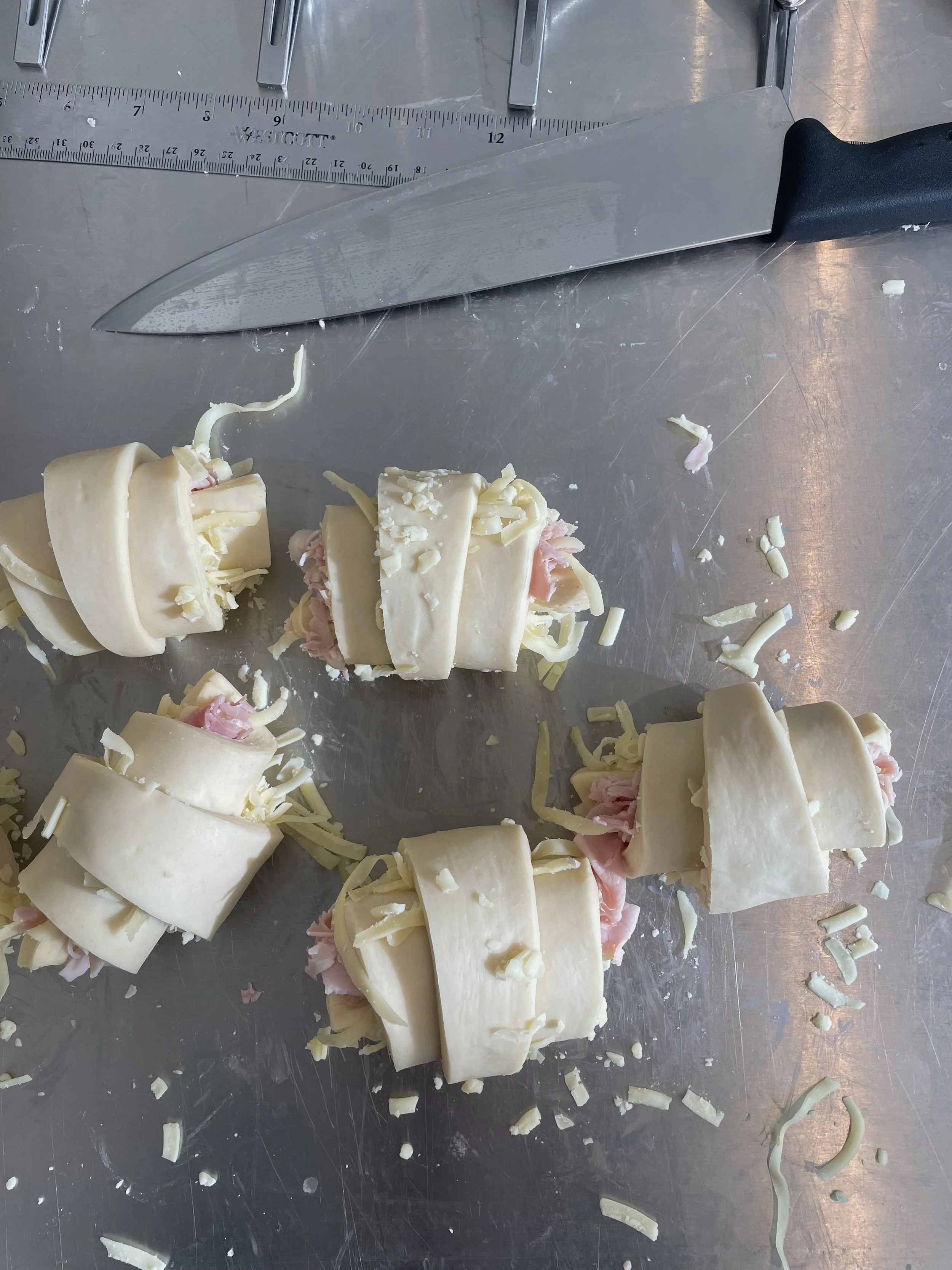 Rolled pastries with cheese and ham on a stainless steel work surface, with a large kitchen knife and a ruler visible at the top.