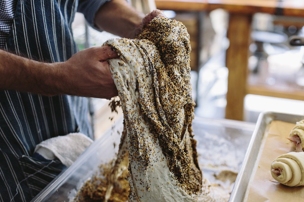 Person wearing a striped apron handling a large piece of dough coated with seeds, likely sesame, in a bakery or food preparation area.
