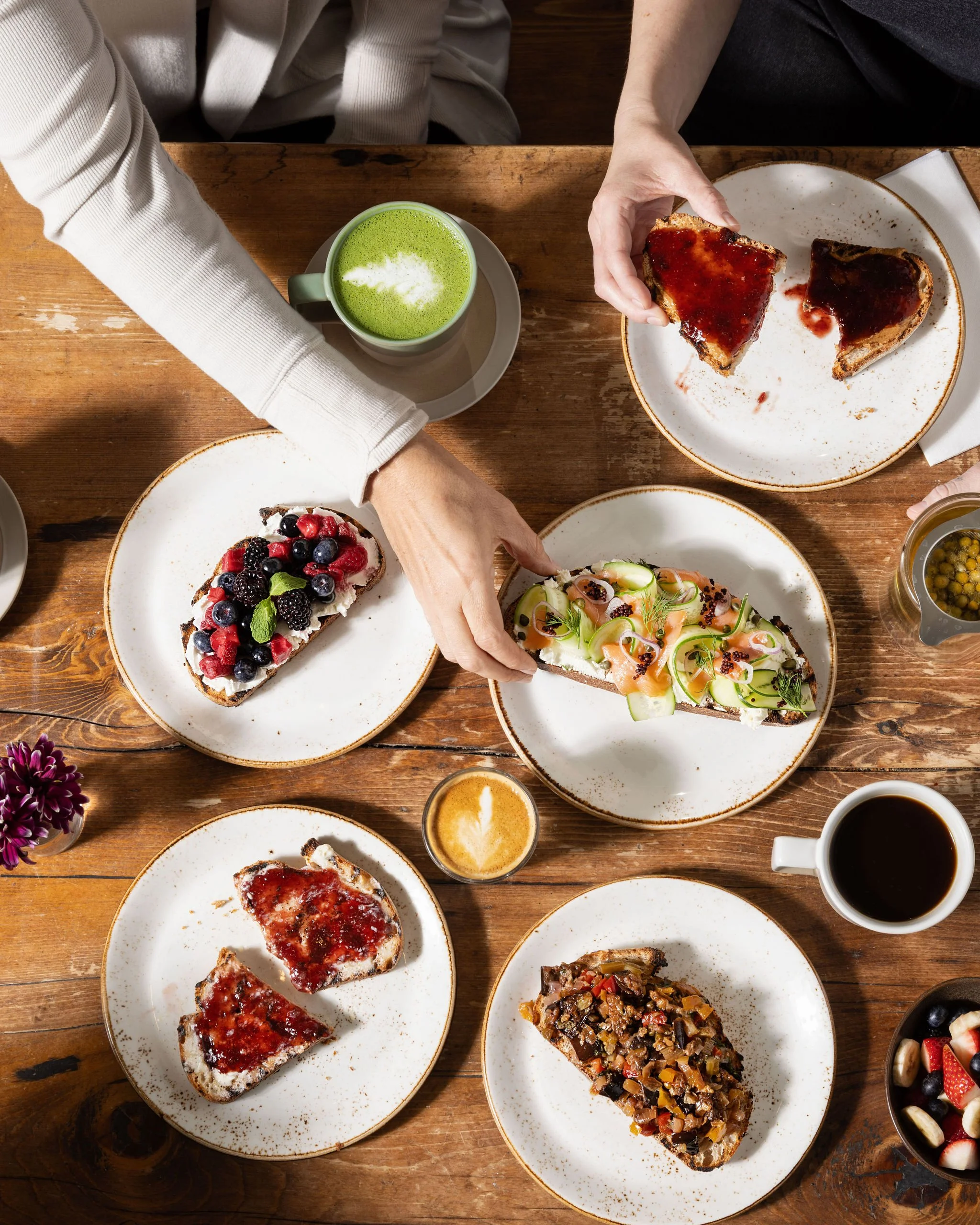 Top-down view of a breakfast table with plates of toast, some with fruit toppings, and others with jam or savory toppings, along with drinks including coffee, a green matcha latte, and a small cup of coffee with latte art.