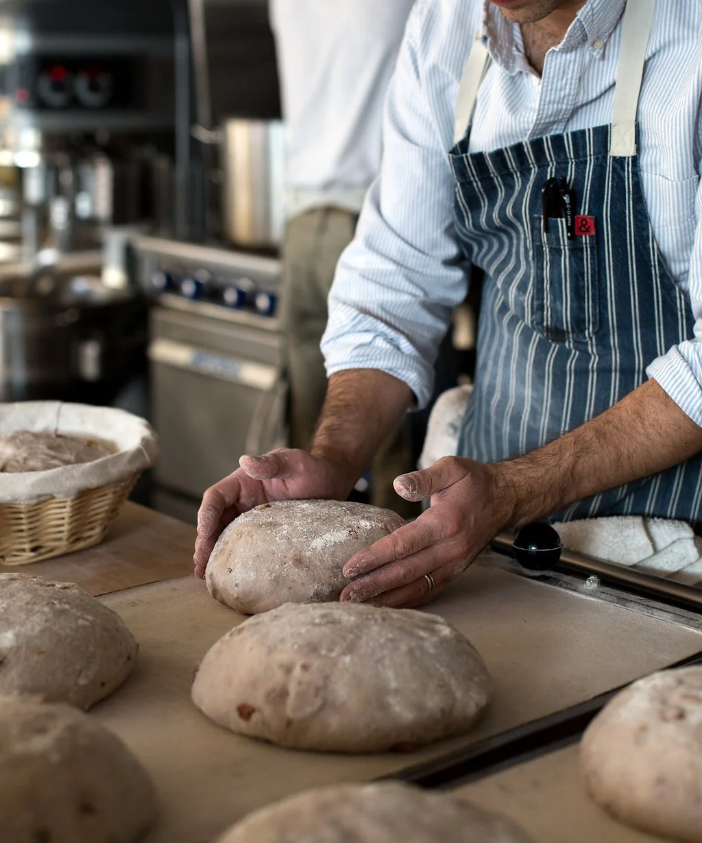 A baker in a striped apron shaping bread dough at a bakery counter with baskets and ovens in the background.