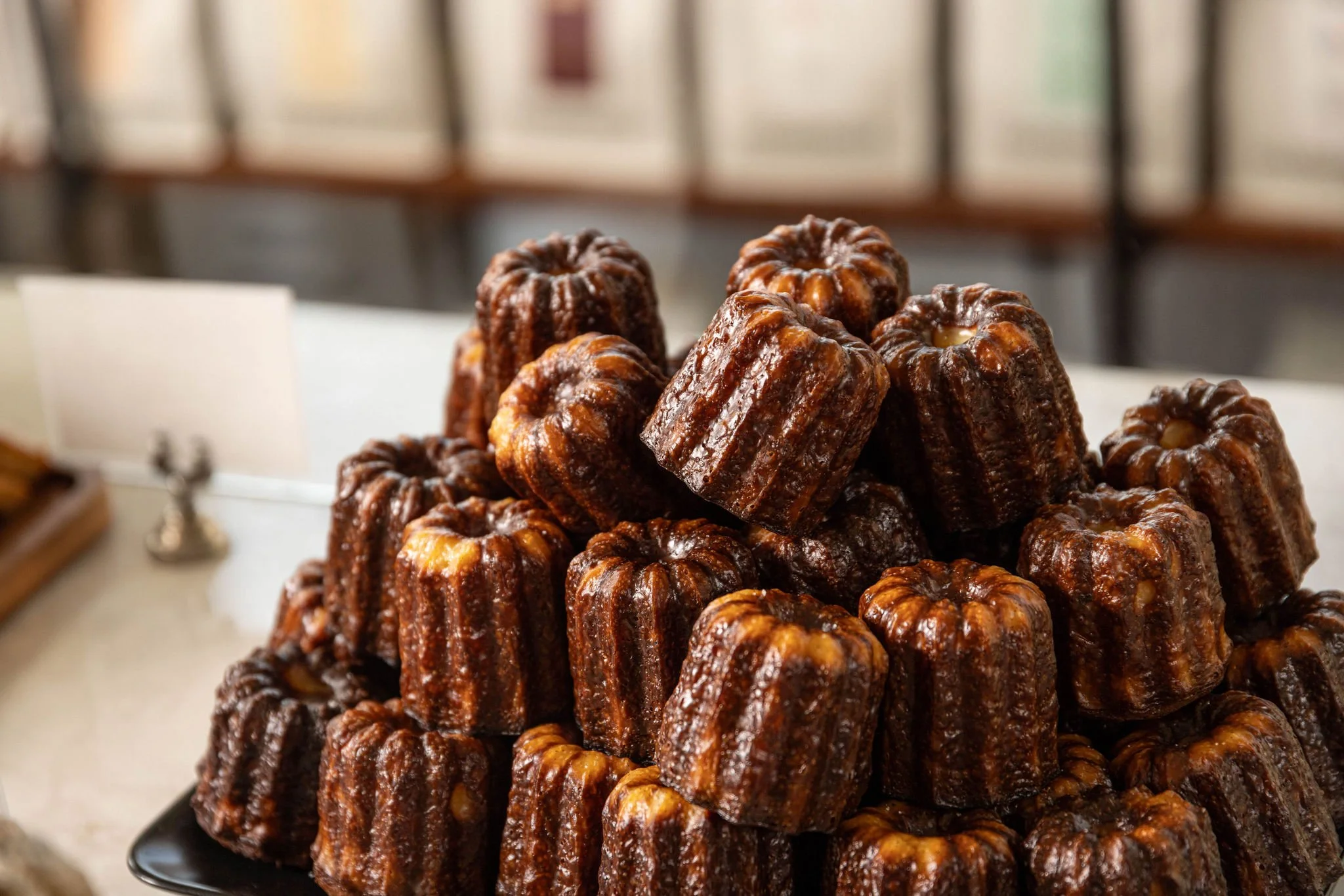 A pyramid of cooked cannelés with a dark caramelized exterior on a black plate in a bakery or shop setting.