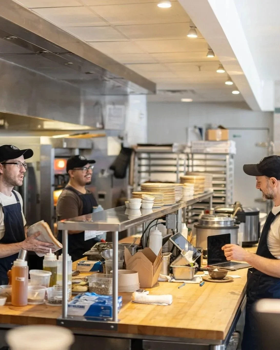 A busy restaurant kitchen with three male chefs wearing black caps and aprons, preparing food at a countertop with stacks of plates, bowls, and condiments.