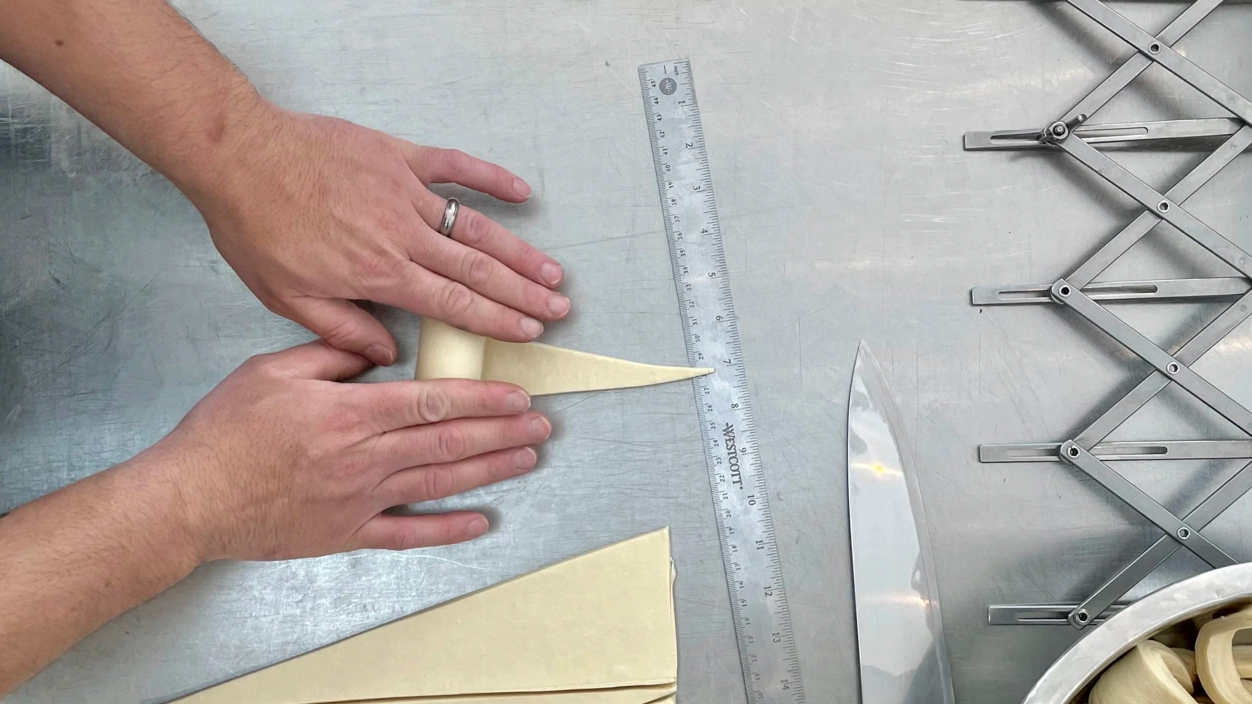 Person folding and shaping pastry dough on a stainless steel surface with a metal ruler, a knife, and a folding metal ruler nearby.