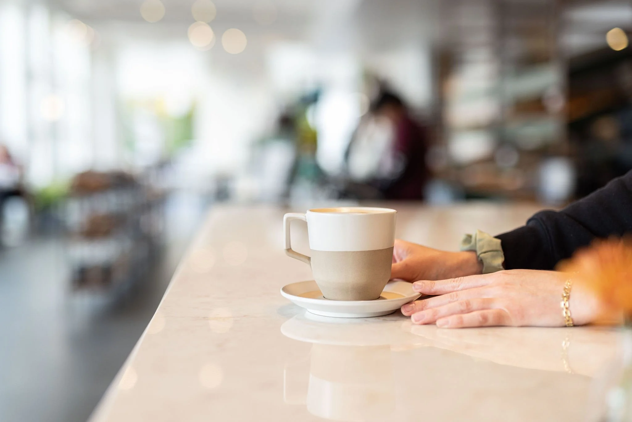 Person's hands resting on a marble counter next to a coffee mug on an espresso saucer in a bright coffee shop.