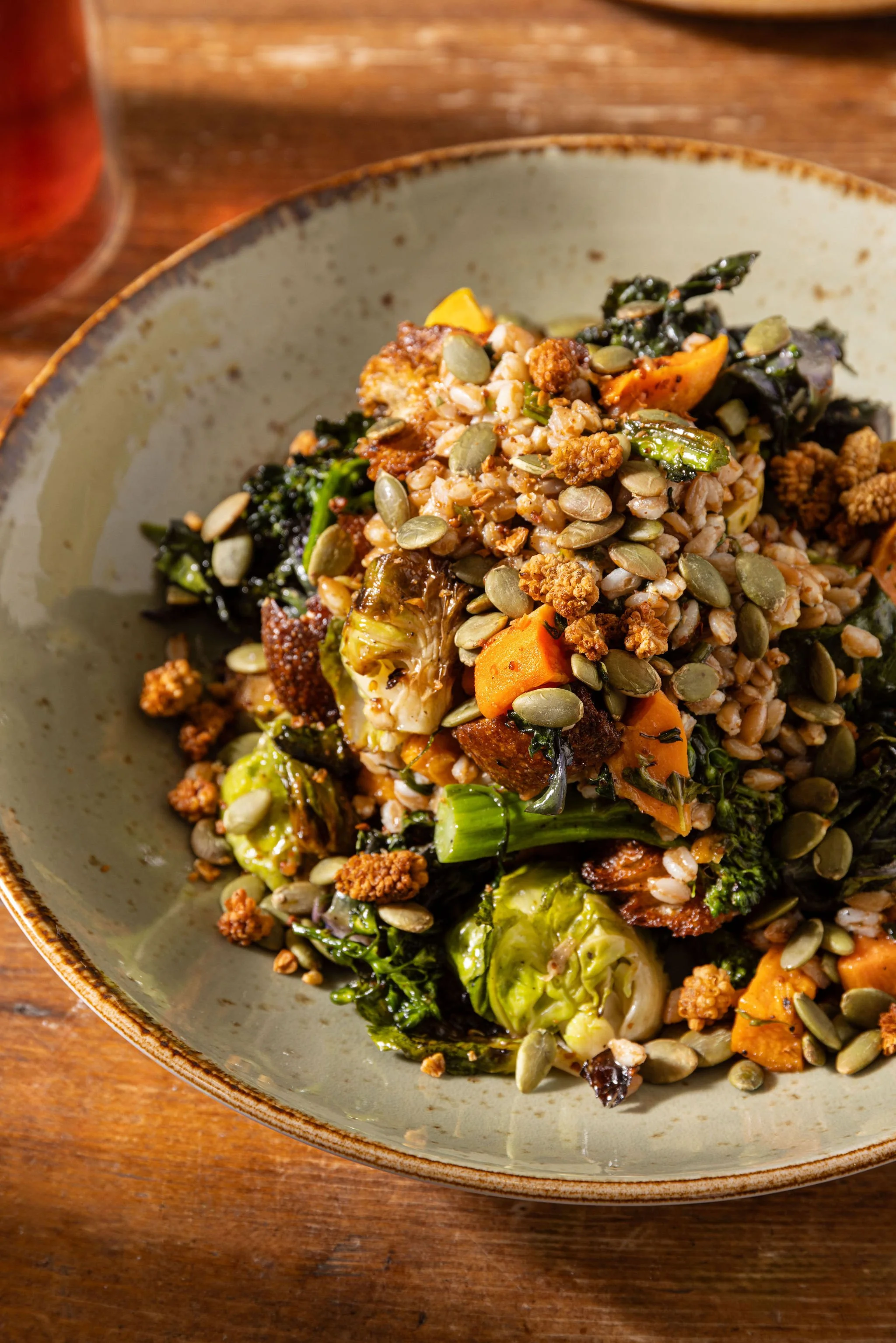 A bowl of roasted vegetable salad with greens, seeds, and grains on a wooden table.