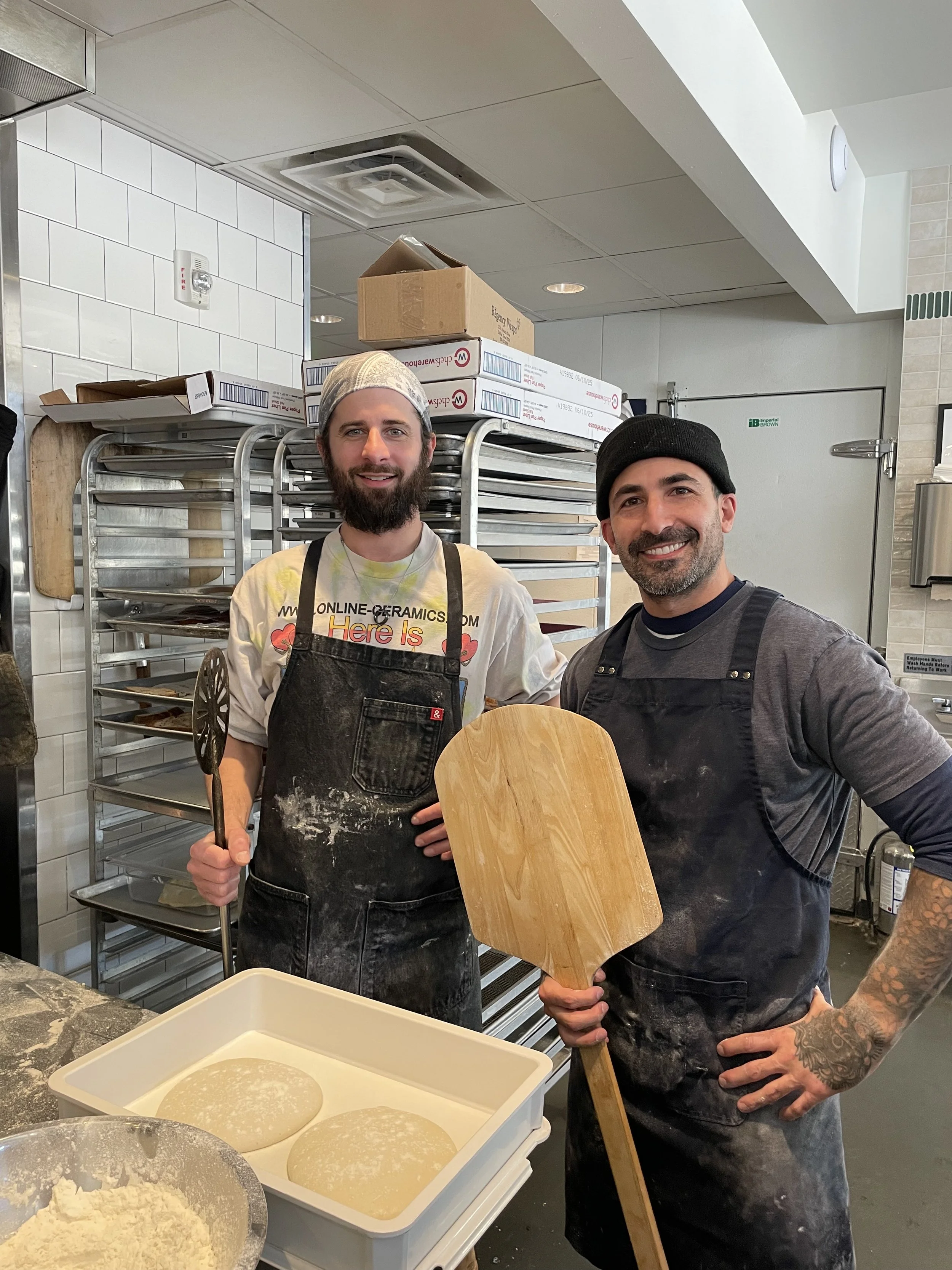 Two men in chef aprons and hats in a bakery kitchen, standing behind dough and pizza pans, holding a wooden pizza peel and a whisk, smiling at the camera.