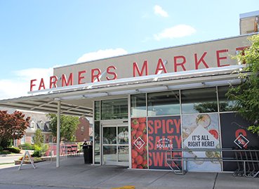 Exterior of a Farmers Market with a sign reading 'Farmers Market' in red letters, large windows, and a banner advertising spicy square.