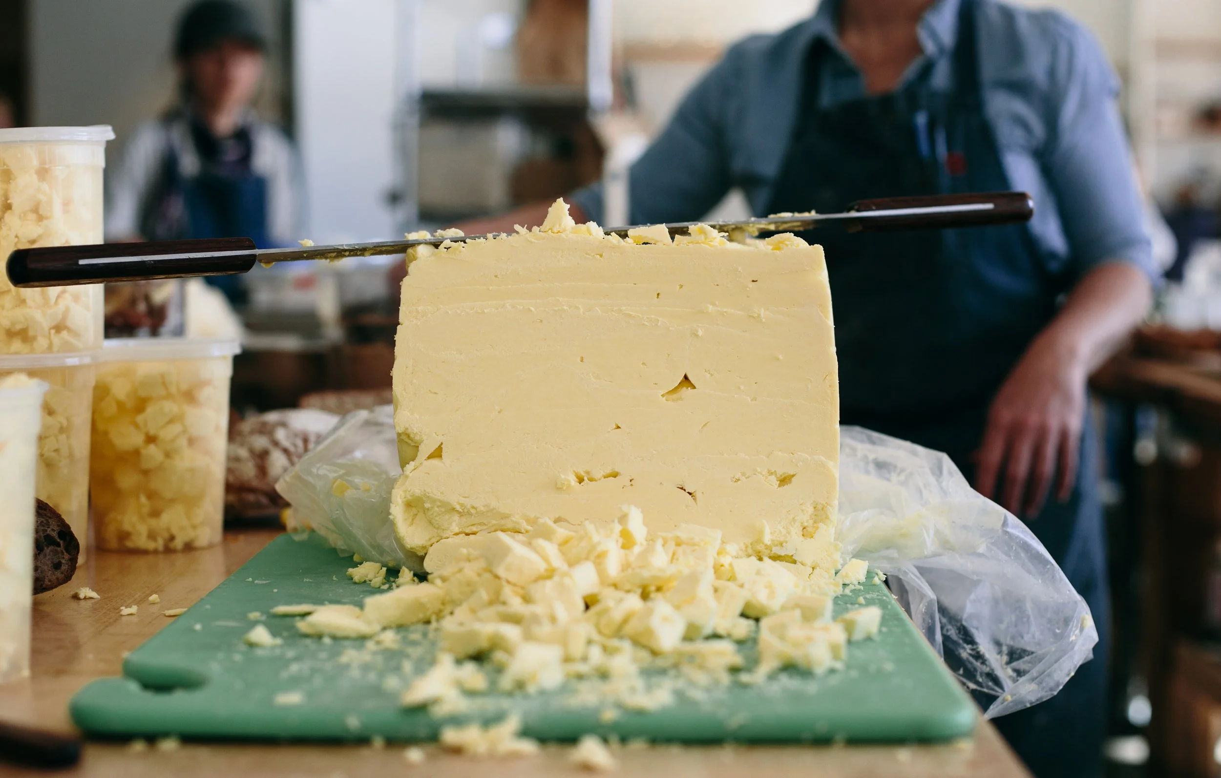 Block of yellow cheese on a green cutting board with cheese shavings, with a person in a blue shirt and apron in the background, and food containers on the side.