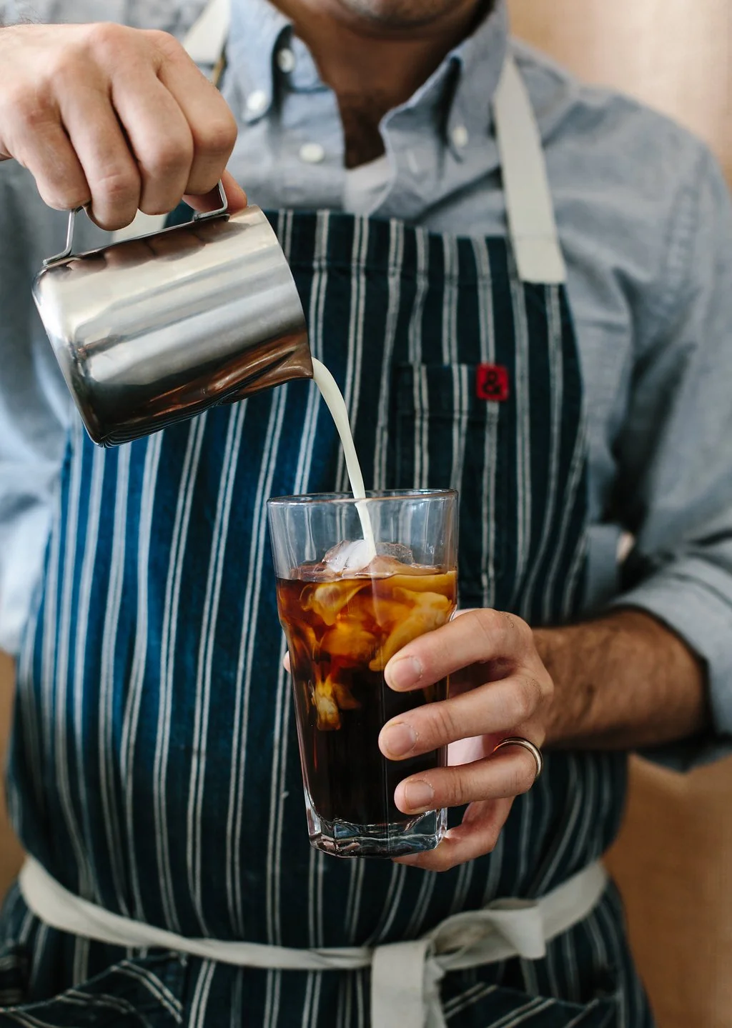A person in a striped apron is pouring cream into a glass of iced coffee.