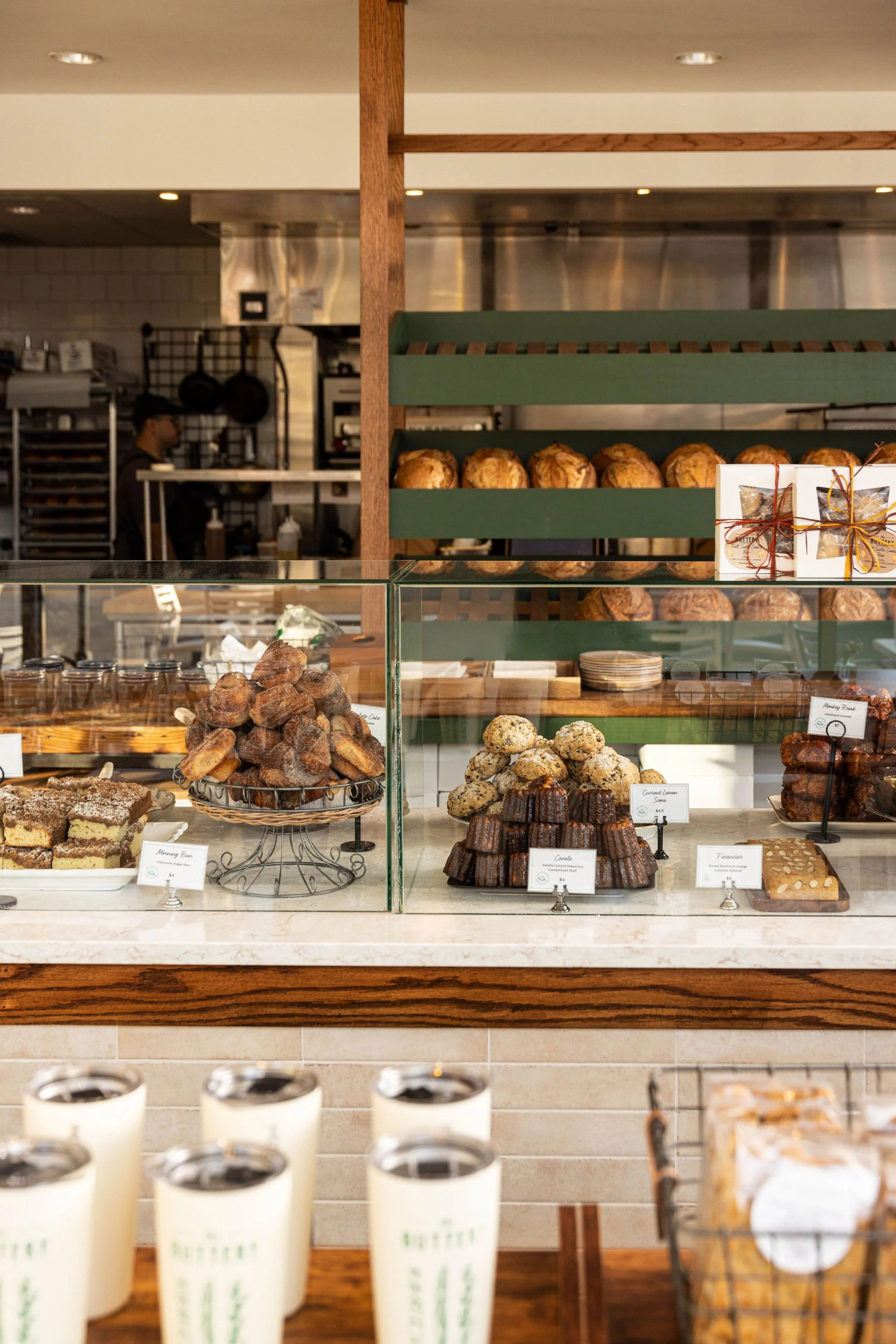 Display case with assorted baked goods, including cookies, bars, and croissants, in a café. In the background, a bakery kitchen with staff, shelves, and equipment are visible.