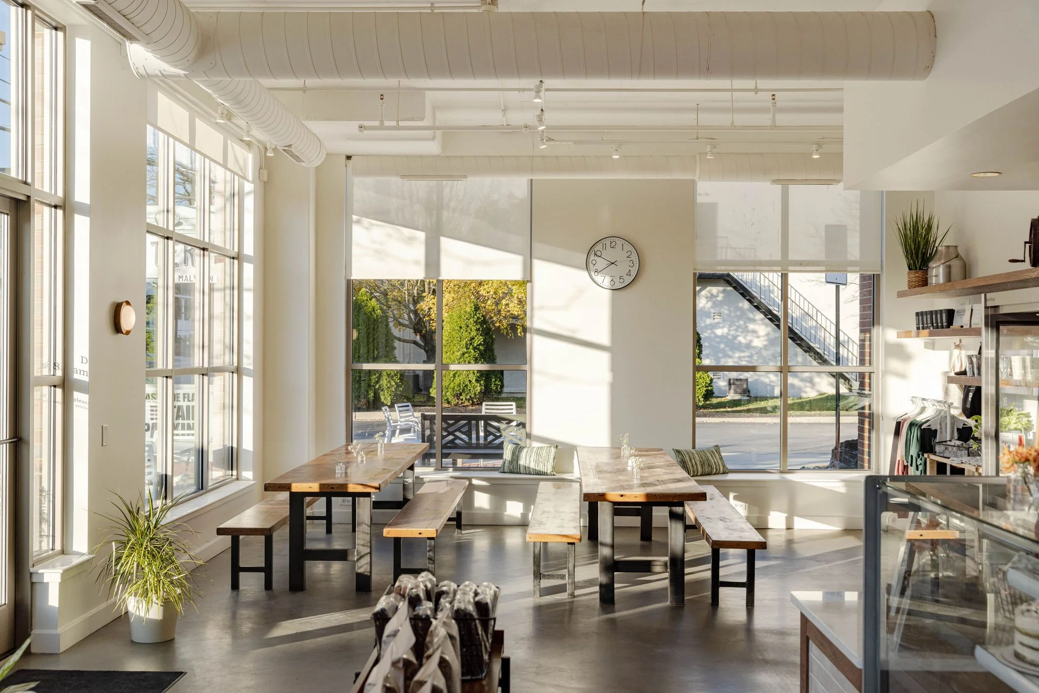 Interior of a bright café with large windows, wooden tables and benches, a wall clock, and outdoor seating visible through the windows.