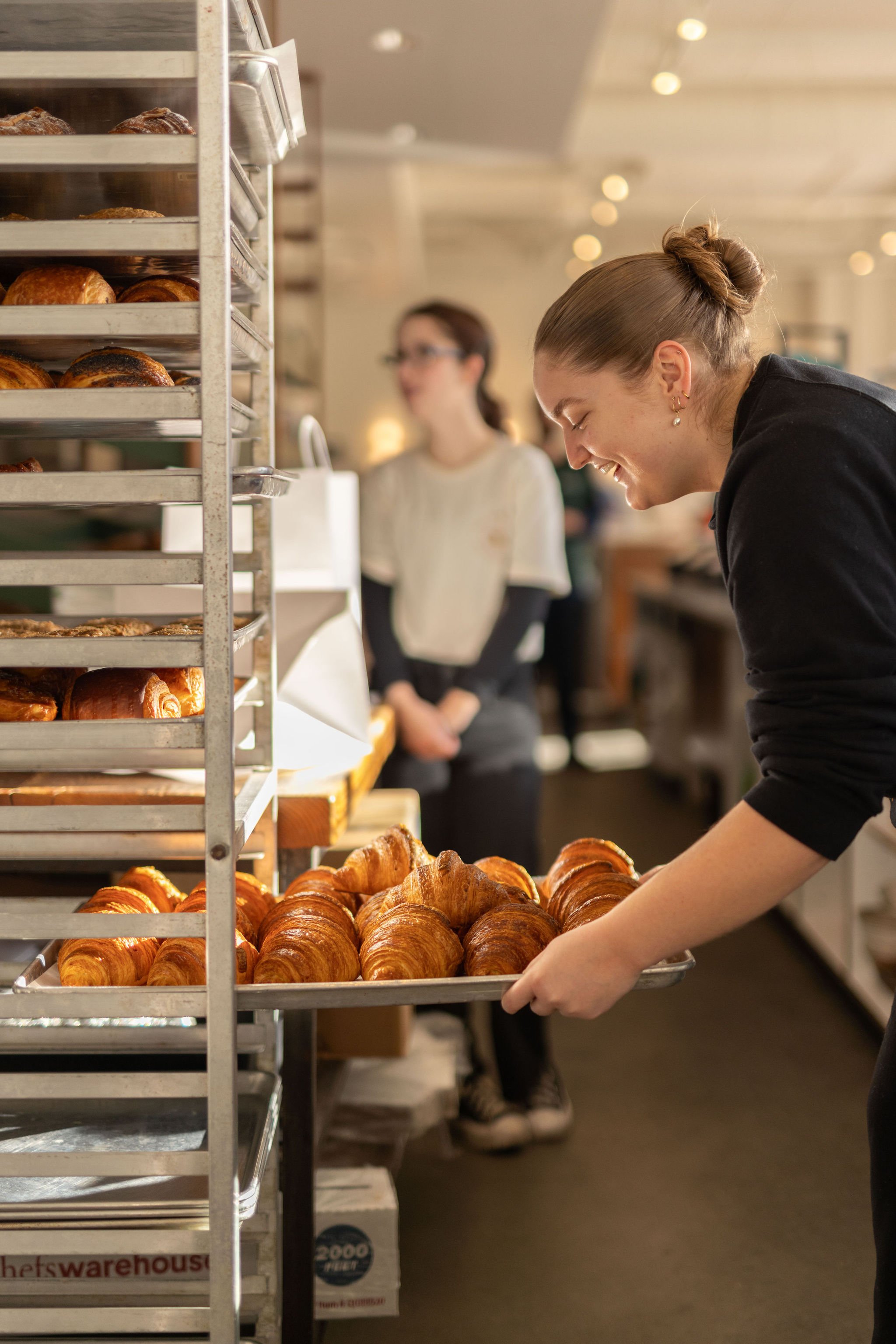 A woman in a black long sleeve shirt and earrings, smiling, is placing a tray of croissants into a bakery display. Behind her, another woman in a white top and glasses is standing near shelves filled with baked goods, in a bakery or cafe setting.