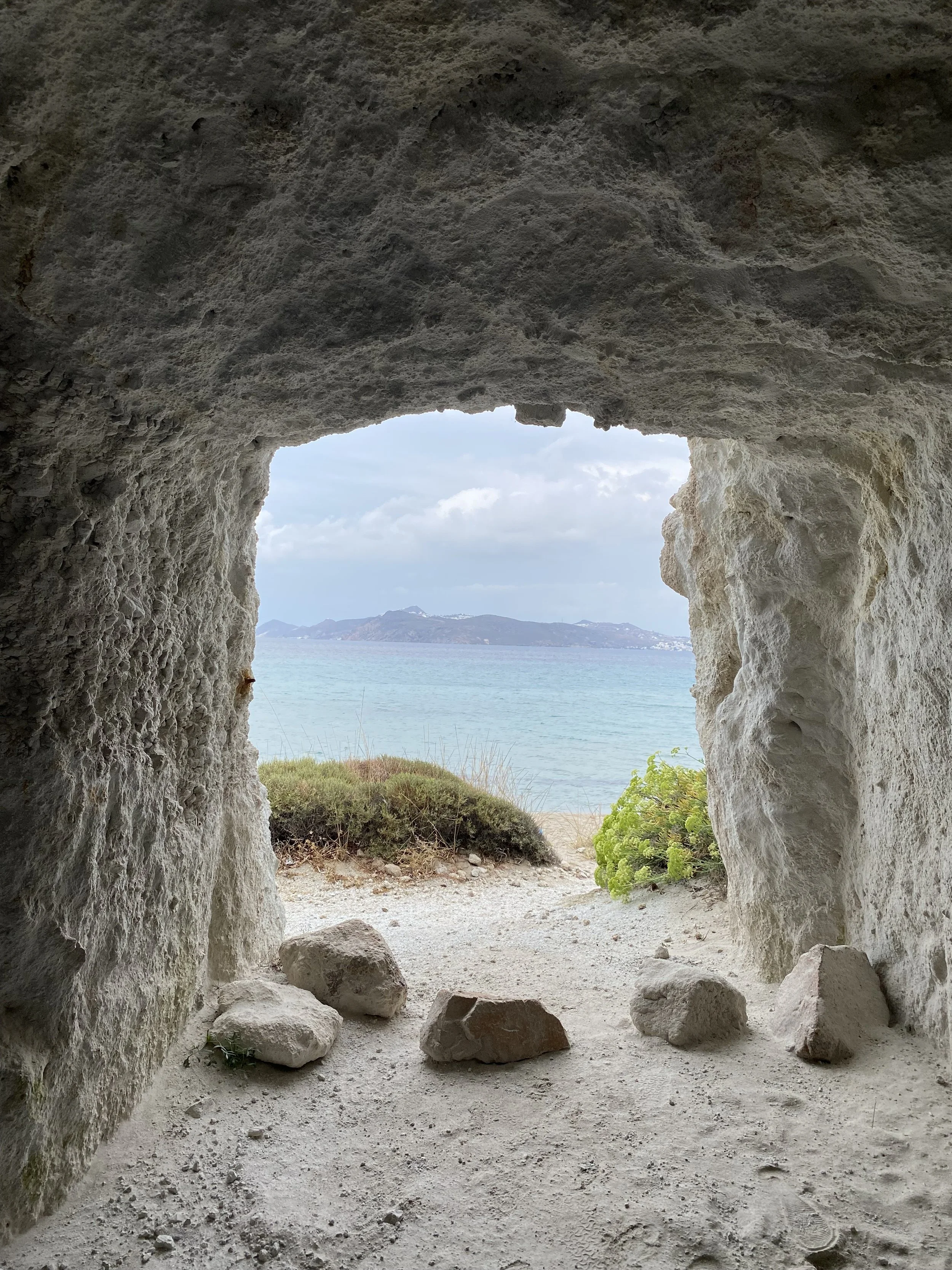 Vue d'une plage vue à travers une ouverture dans une grotte en pierre avec des rochers au sol, des buissons, et la mer avec des îles au loin, sous un ciel partiellement nuageux.